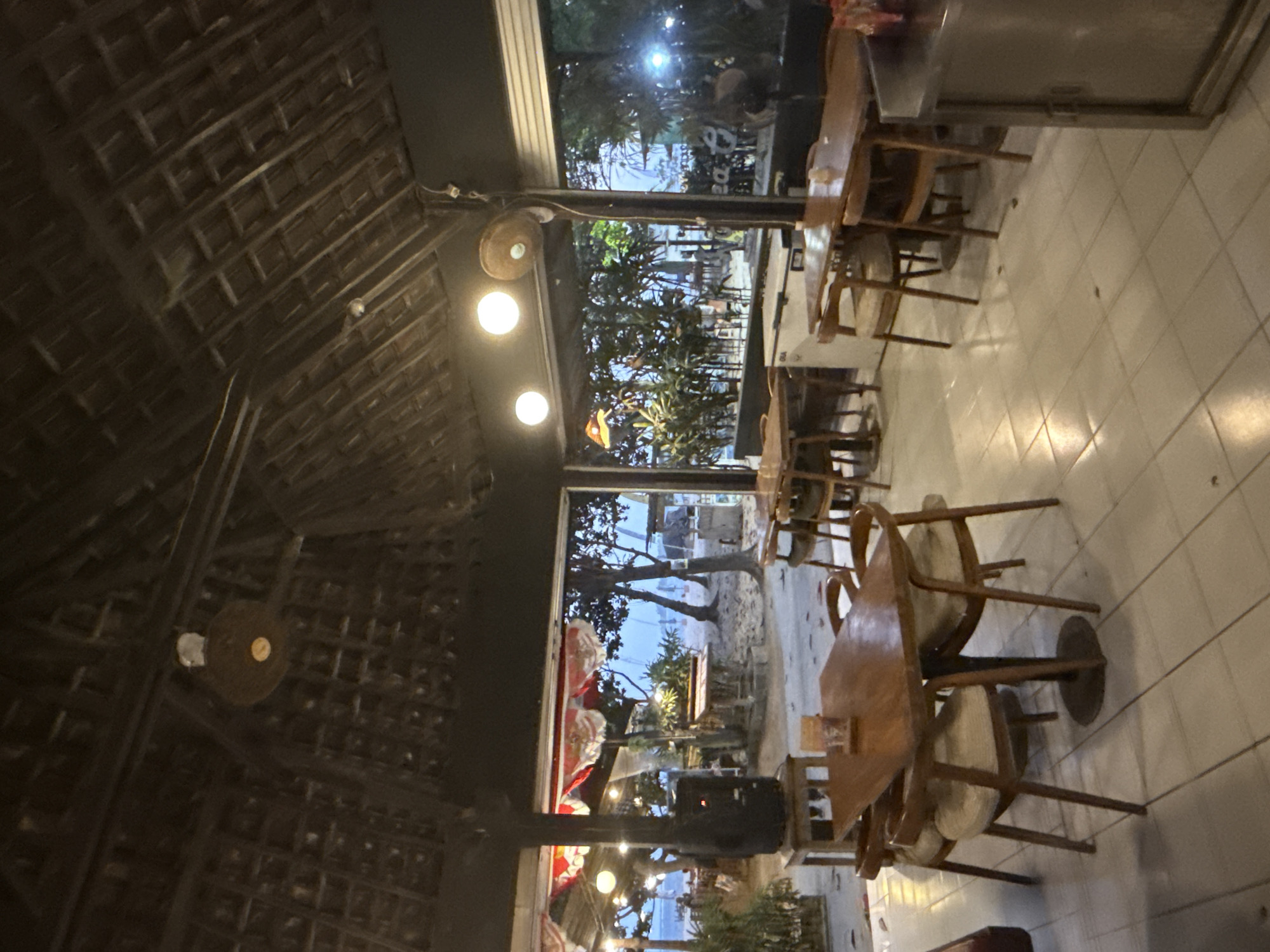 The image depicts the interior of a casual open-air restaurant with a thatched roof, likely located on a beach in Denpasar, Indonesia. The restaurant appears to be near the coast, as evidenced by the sandy area visible through the open front, with palm trees and other vegetation in the background. Beach umbrellas and what appear to be small huts are also visible on the sand.
Inside the restaurant, several wooden tables with chairs are arranged on a tiled floor. The tables are simple, with wooden tops and dark-colored bases. The chairs are also wooden, with upholstered seats. Some tables have small decorative items or menus on them.
In the background, through the open facade of the restaurant, several more tables and chairs are set up on the sand, suggesting a larger seating area. A large black speaker sits on a wooden stand to the left, and a white refrigerator or freezer unit is visible behind a table on the right.
Overhead, a rustic thatched roof is supported by wooden beams. Light fixtures, including what appear to be spherical lamps and a ceiling fan, are mounted to the ceiling. The lighting is warm and ambient, creating a relaxed atmosphere. The overall impression is that of a beachside dining establishment, likely popular for its casual ambiance and proximity to the ocean. There are no visible people in the foreground or midground of the image, though there are faint figures and activity visible on the beach in the distance.