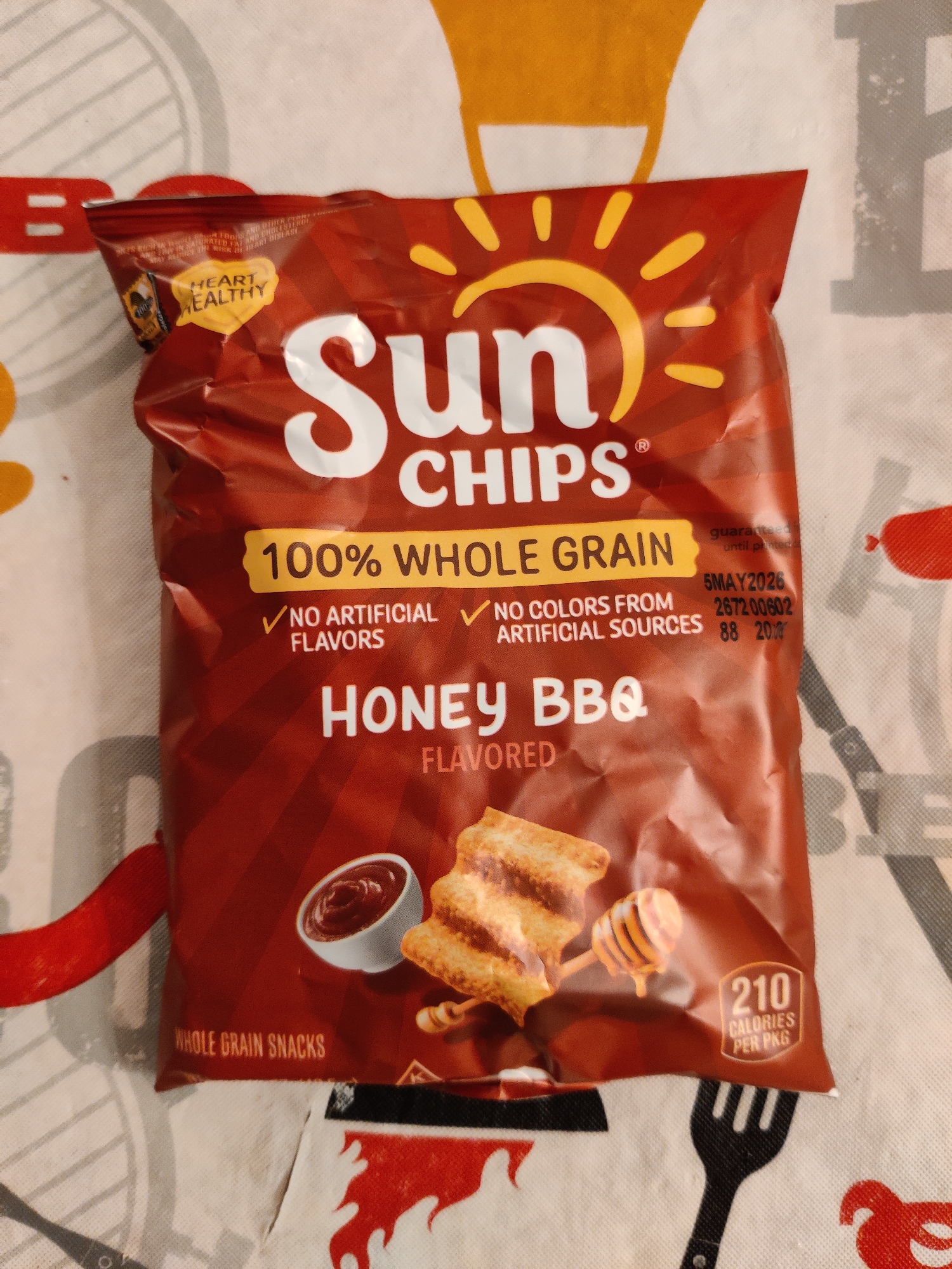 A SunChips Honey BBQ bag is lying on a patterned tablecloth. The bag is primarily a dark red color with a yellow sun logo and white text. Text on the bag indicates "100% WHOLE GRAIN," "NO ARTIFICIAL FLAVORS," and "NO COLORS FROM ARTIFICIAL SOURCES." The flavor "HONEY BBQ" is prominently displayed. There are images of chips, a bowl of sauce, and a honey dipper with honey dripping. A nutrition label shows "210 CALORIES PER PKG." In the top left corner of the bag, a "HEART HEALTHY" seal is visible. The background is a white and gray patterned fabric. The image does not provide any information about the location in Lower Paxton Township, United States of America.