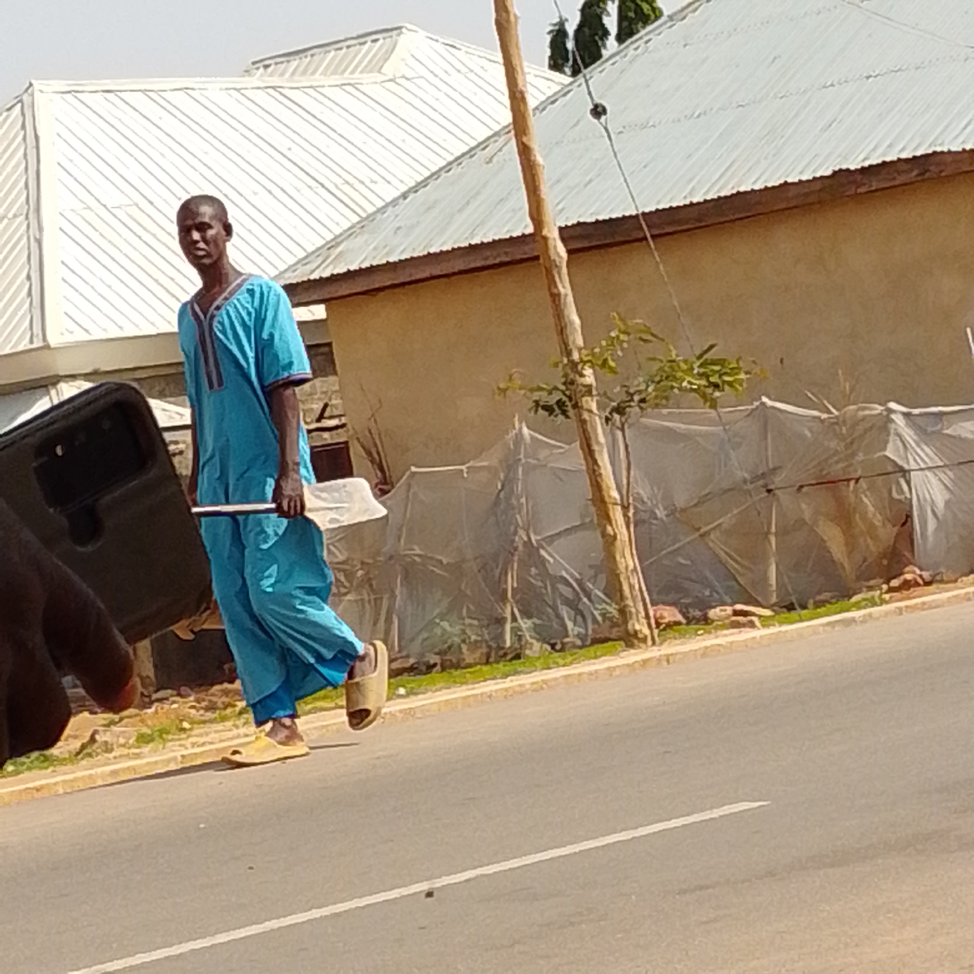 A man in a light blue traditional outfit and yellow sandals walks on a paved road in front of a building with a corrugated metal roof. He is holding a long, narrow, white object, possibly a tool or equipment, horizontally. To his left, a dark object, potentially a mobile phone, is being held by a person's hand. In the background, the building has tan walls, and some structures covered in translucent material are visible behind a wooden pole. The scene appears to be in Gombe, Nigeria.