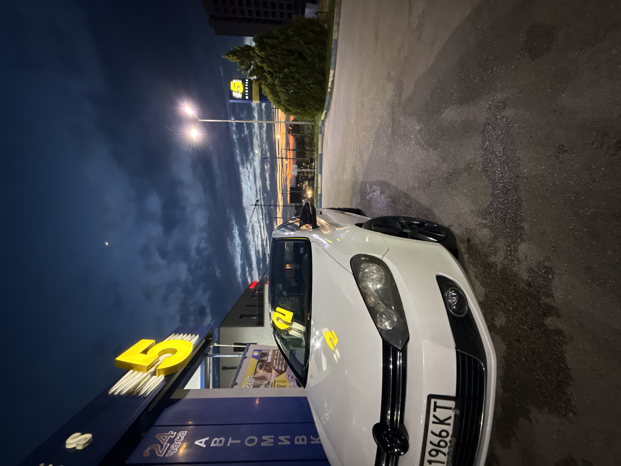 The image depicts a white Volkswagen Golf parked at what appears to be a car wash or commercial establishment in Pleven, Bulgaria, during dusk or early night. The car is prominently featured in the foreground, angled slightly to the right, showcasing its front end, black grille, and black alloy wheels. Yellow decals, possibly the number '2' or '5', are visible on its windshield and driver's side window. The Bulgarian license plate "1966 KT" is clearly visible on the front bumper.
On the left side of the frame, a building features a large, illuminated yellow number '5' sign. Below this, Cyrillic text reads "АВТОМИВКА" (Avtomivka), which translates to "car wash," and further below, "24 часа" (24 chasa), indicating 24-hour operation. Streetlights illuminate the scene, creating bright flares.
The sky is a notable element, dominated by dark blue and grey clouds, with a clear crescent moon visible in the upper left. A striking band of orange and red light on the horizon suggests either a sunset or sunrise, contrasting with the darker sky. In the middle ground, trees and distant buildings are visible. The ground is dark asphalt, appearing somewhat wet. The overall scene conveys a calm, transitional time of day.
