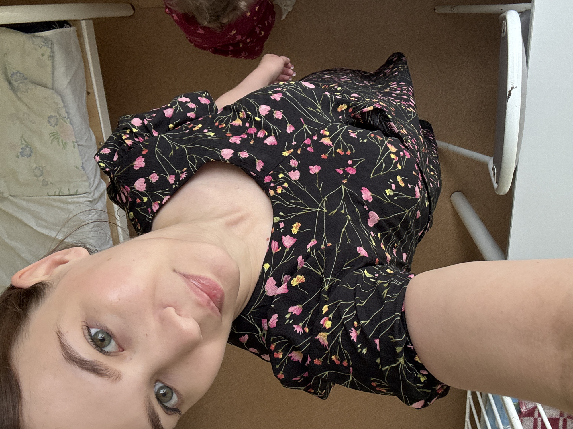 The image is a high-angle selfie of a young woman in a black floral-print dress. She has fair skin, green eyes, and dark brown hair pulled back. She is wearing light makeup. The background appears to be an indoor setting with a wooden floor and a white bed frame. Part of another person, possibly a child, wearing a red garment is visible in the background to the right. There are also some white furniture elements in the foreground. The image does not contain any identifiable landmarks or elements that would indicate its location in Chaikovsky, Russia.