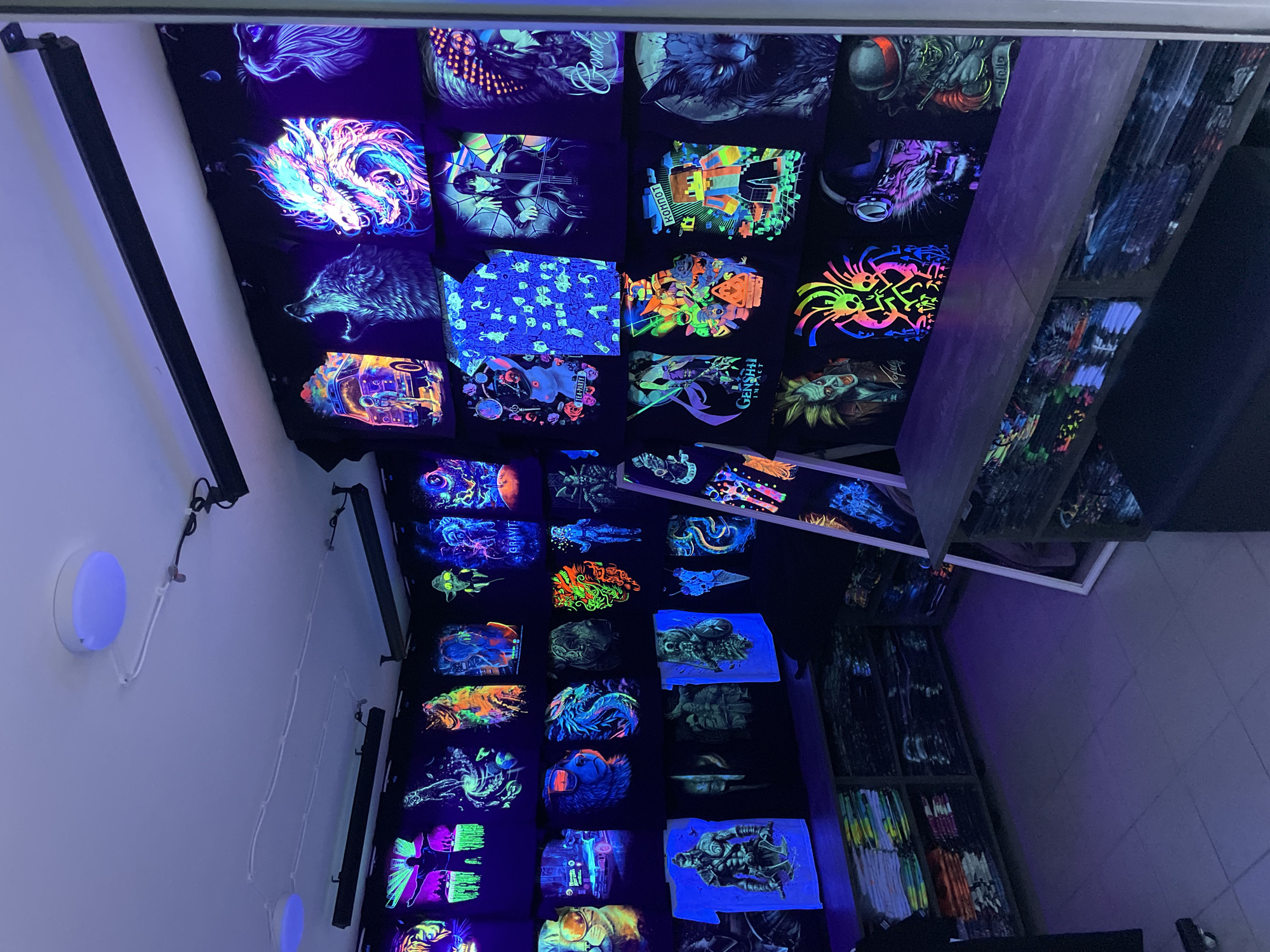 A brightly lit merchandise display with black t-shirts that have neon or fluorescent designs is visible. The shirts are hanging on racks or shelves. A shelving unit is placed on the right filled with folded t-shirts. There are lighting fixtures mounted on the wall, casting a purple or UV glow over the scene. The merchandise display is located in Sochi, Russia.