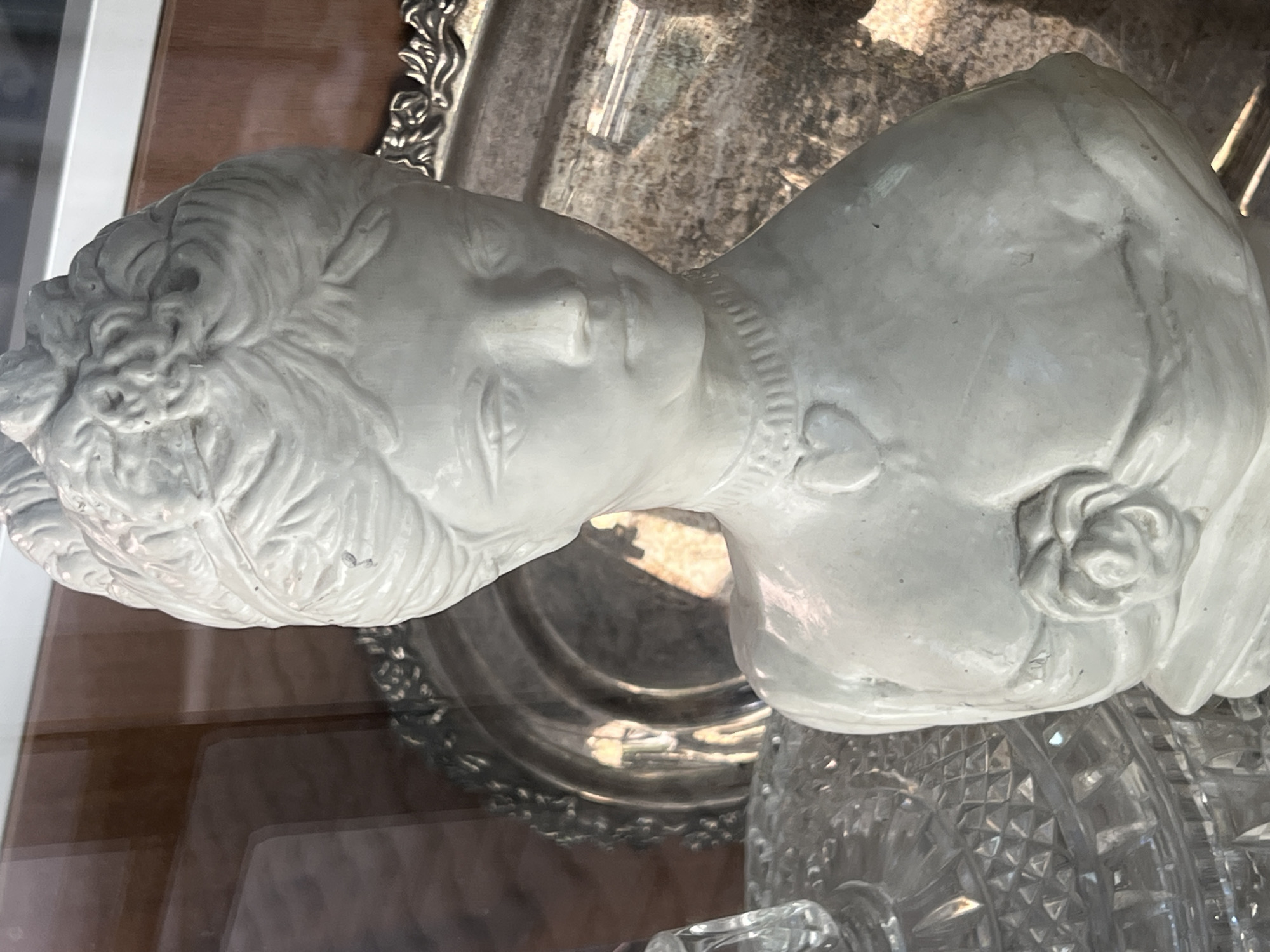 The image shows a plaster bust of a young woman with an ornate hairstyle, a necklace with a heart pendant, and a rose at her chest. The bust is white and appears to be a decorative item. It is placed in front of a reflective surface, possibly a glass cabinet, behind which a silver platter with an embossed rim is visible. To the left of the bust, a portion of a cut-glass object, likely a vase or decanter, is visible. The background suggests an indoor setting with shelves and display items, hinting at a shop or a collector's display. There is no indication of a specific location within Sofia, Bulgaria.