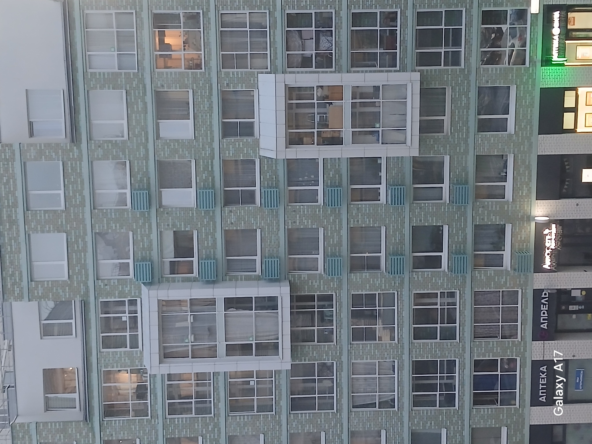 The image depicts the facade of a building in Yekaterinburg, Russia. The building is constructed with a light green brick facade, punctuated by numerous windows arranged in a grid pattern. Several windows are grouped together to form balconies. The right edge of the image shows a portion of an adjacent building with signage in Cyrillic script including the words "АПРЕЛЬ" and "АПТЕКА". The phrase "Galaxy A17" is also visible on the right side of the image. The sky is overcast.