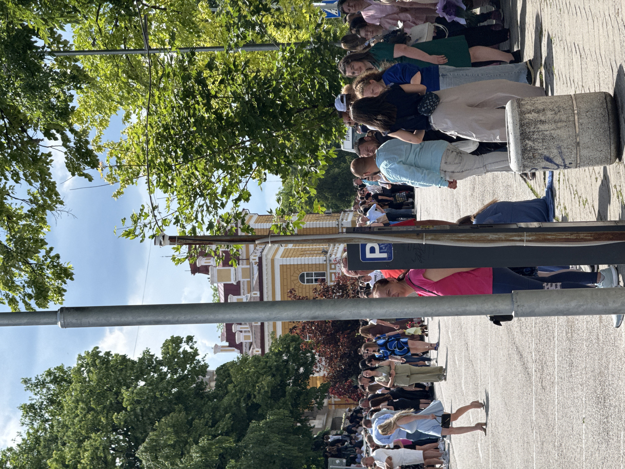 The image depicts a street in Shumen, Bulgaria, with a crowd of people gathered on the sidewalk. A yellow building with a red roof is visible in the background. The image also includes trees, a blue parking sign, and a metal pole. The people appear to be standing and observing something out of frame, possibly a performance or event. The image is oriented vertically.