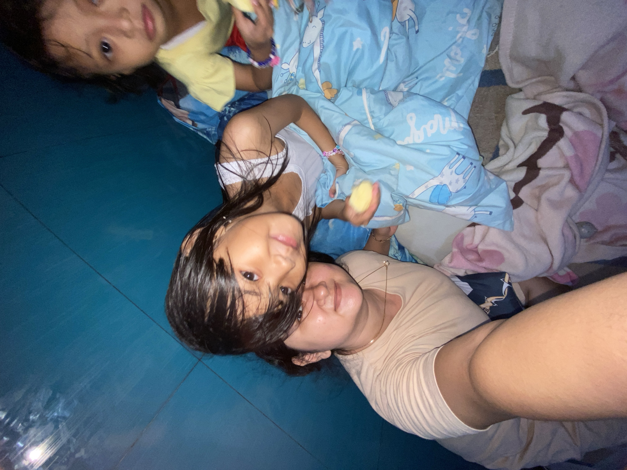 Three individuals are visible in an indoor environment, likely a domestic residence in Gelam, Indonesia. A woman with short, dark hair and fair skin is taking a selfie while lying down on a blue, patterned bedspread with cartoon motifs. A young girl with long, dark hair is lying on top of the woman, looking at the camera and holding a piece of food. Another young girl is partially visible in the top left corner of the frame, also holding food and looking toward the camera. The floor is a dark blue color. A pink and white blanket is visible to the right of the bedspread.