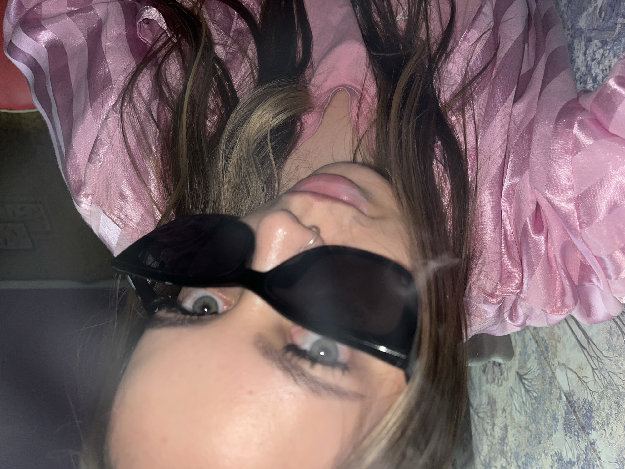 A person with long brown hair and sunglasses lies on their back, wearing a pink striped top. They have a nose ring. The background shows a patterned surface. The location is unspecified in the image and cannot be determined as Novopokrovskaya, Russia.