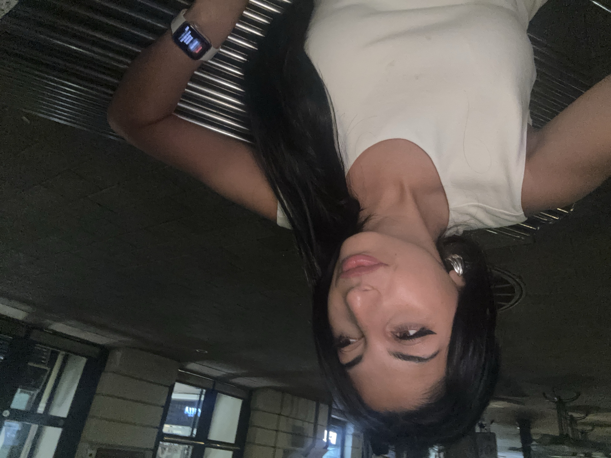 A medium shot shows a woman sitting on a metallic bench outdoors at night, with buildings and a street visible in the background. The woman has long, dark hair, brown eyes, and is wearing a white sleeveless top and large hoop earrings. She is looking to her right with her mouth slightly open. A white smartwatch is visible on her left wrist. The scene appears to be in an urban environment, likely a city, but the specific location of Plovdiv, Bulgaria cannot be determined from the image alone.