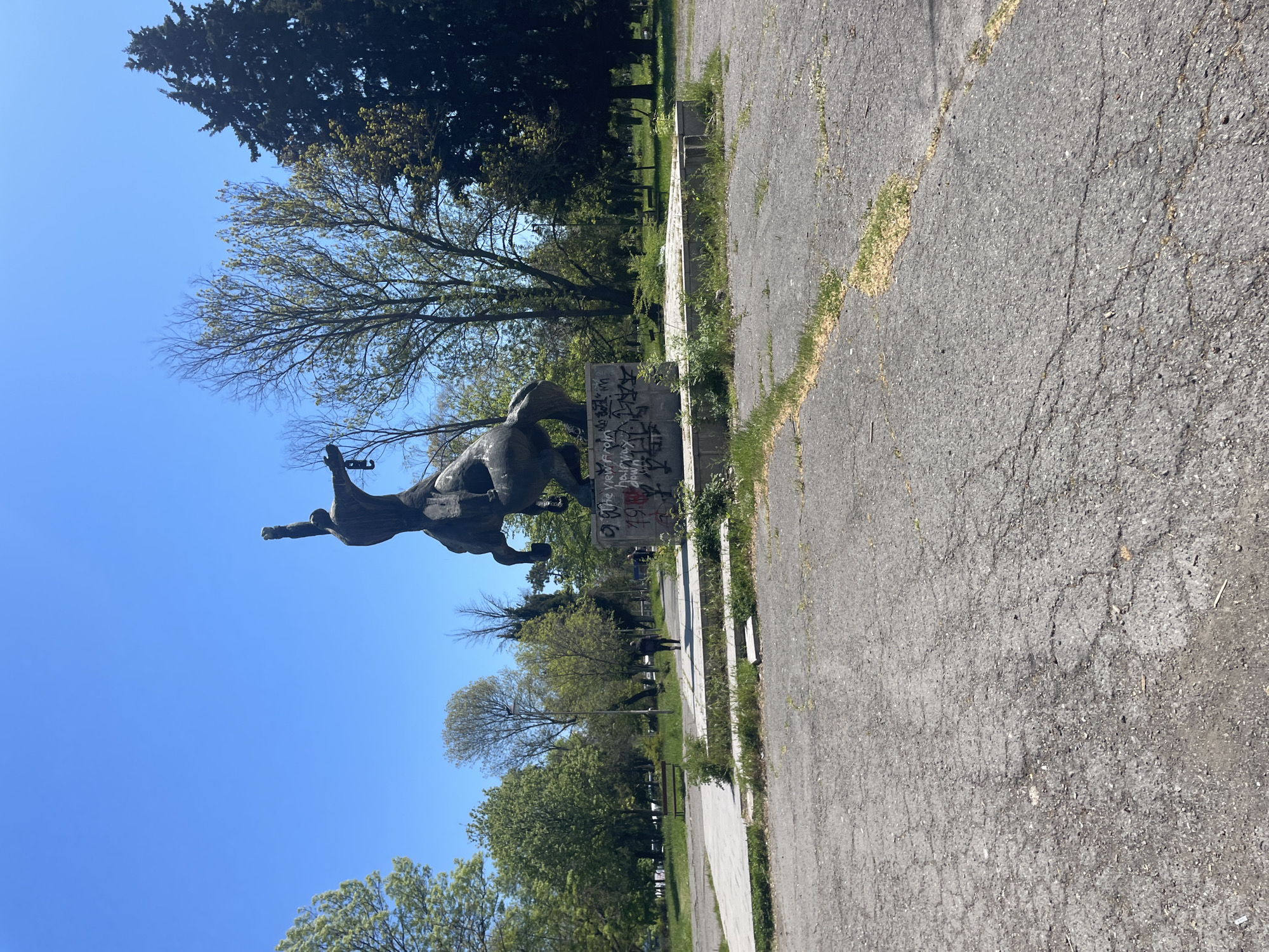 The image depicts a bronze statue of a male figure riding a horse, located in a park. The statue is mounted on a concrete base with graffiti. The background includes green trees and a clear blue sky. The foreground shows a cracked asphalt pavement with weeds growing along the edges. In the distance, there are other park elements like benches. The statue is the monument of Alyosha, located in Burgas, Bulgaria. Two individuals are seen walking in the distance.