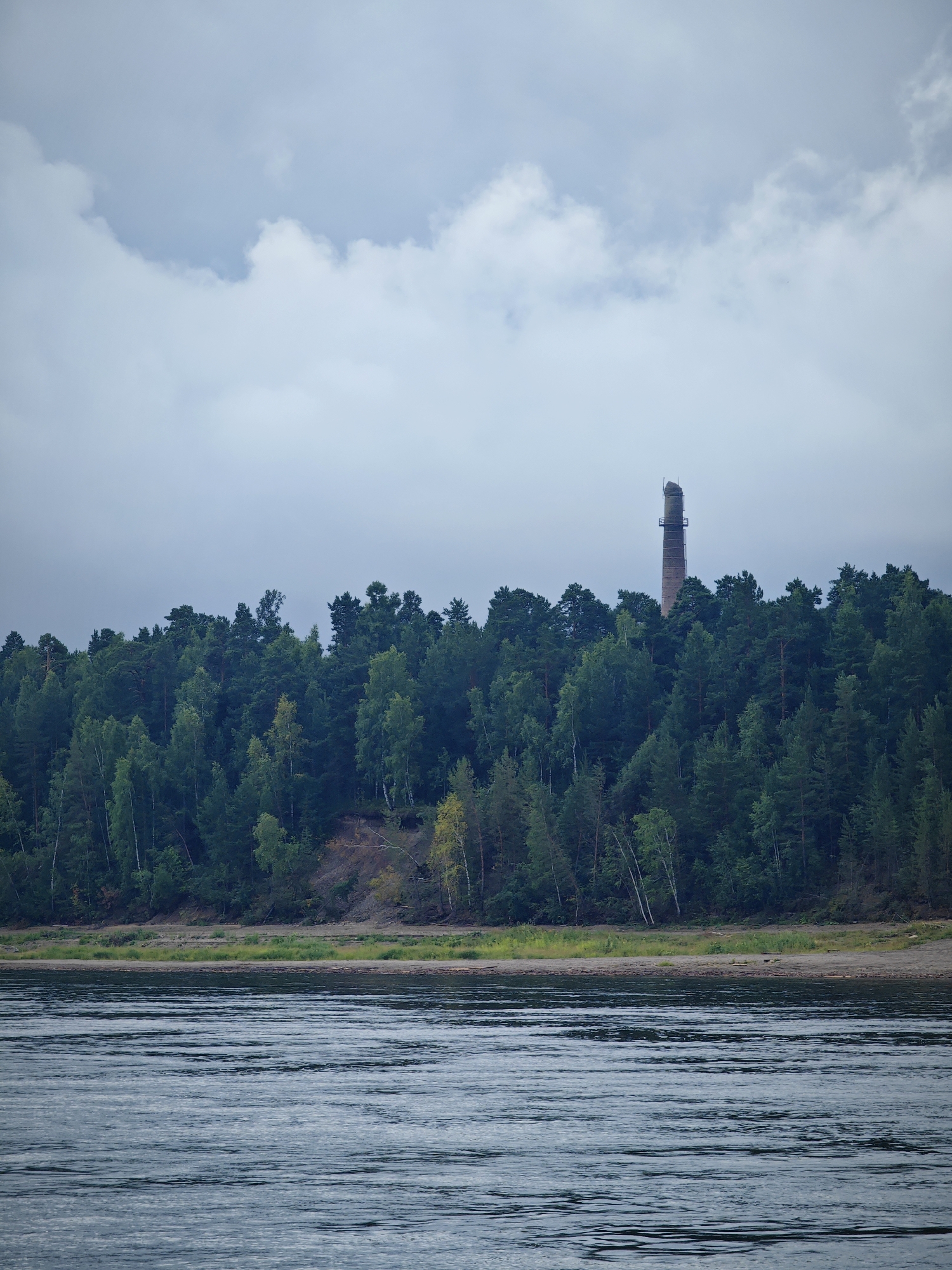 The image depicts a wide, calm river in the foreground, with gentle ripples on its surface. Beyond the river, a sandy bank with sparse vegetation leads up to a dense, green forest. The forest covers a hillside, with various shades of green indicating different tree species and their varying heights. Prominently visible above the treeline, towards the right side of the frame, is a tall, brick industrial chimney. The sky above is overcast with soft, diffused light, suggesting a cloudy day. The scene is devoid of any people or animals. The overall impression is one of a natural landscape with a solitary industrial element, likely situated in or near the city of Lesosibirsk, Russia, given the context of the prompt.