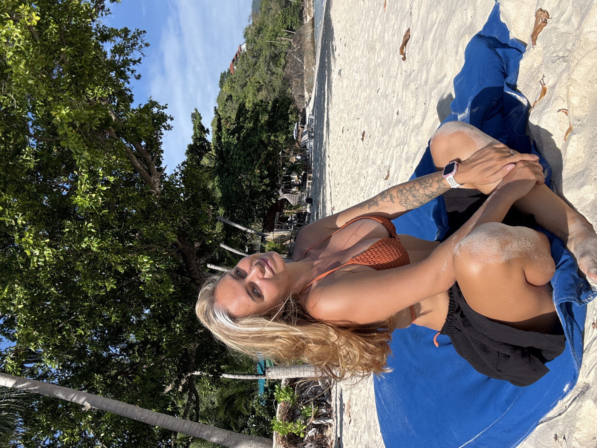 A woman with blonde hair sits on a blue mat on a sandy beach. She is wearing a brown crochet bikini top and a black skirt. Her legs are crossed, and her arms are resting on her knees. She has a tattoo on her left arm and is wearing a white watch. The beach is lined with palm trees and lush green foliage. In the background, the ocean meets a rocky shoreline with buildings on a hillside. The sky is clear and blue. The scene appears to be taken in Ko Samui District, Thailand.