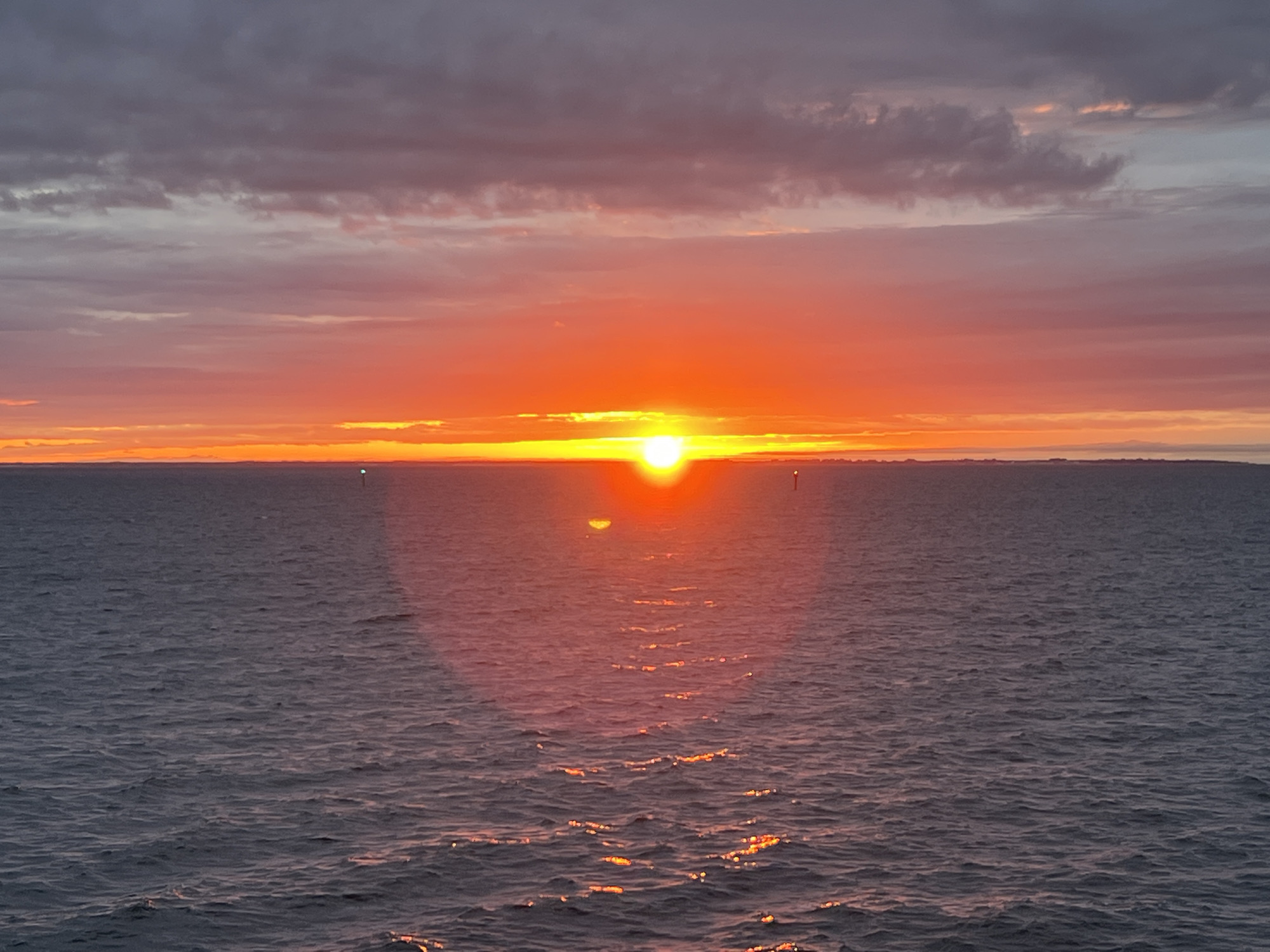 A wide-angle, eye-level, horizontal shot captures a sunset over the Baltic Sea, with the city of Gdynia, Poland, visible as a distant, hazy silhouette on the horizon. The sun, a bright orange orb, is low on the horizon, casting a vibrant glow across the sky and reflecting a shimmering path on the choppy, dark blue water. The sky is a canvas of dramatic colors, transitioning from fiery oranges and reds near the sun to soft pinks and purples, with streaks of dark, gray clouds overhead. Two small, indistinct buoys, one on the left and one on the right side of the frame, are visible in the water. The overall mood is serene and atmospheric, with the vastness of the sea and sky dominating the scene.