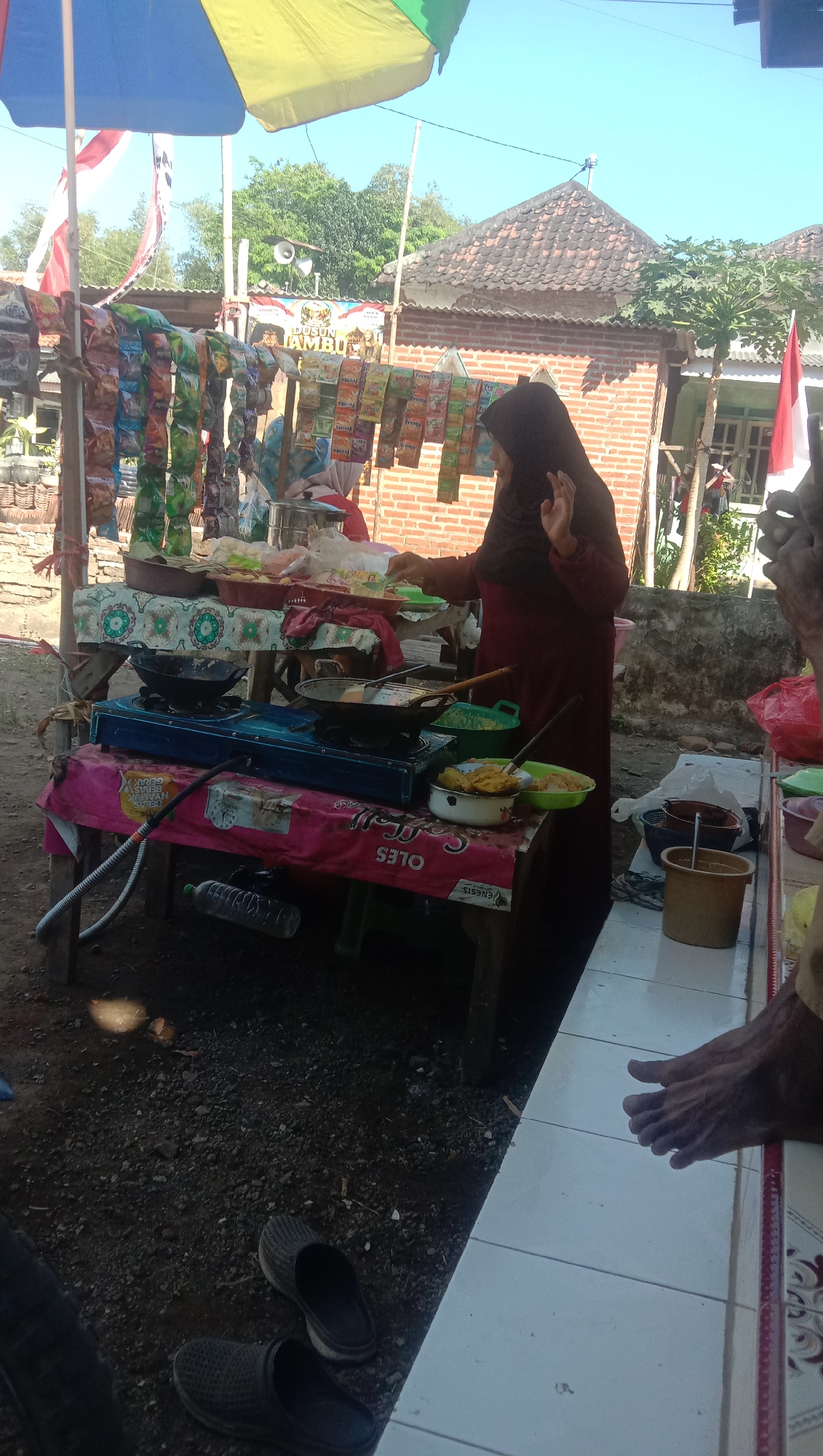 In Ngempit, Indonesia, a woman wearing a dark hijab and a maroon long-sleeved dress is standing behind a food stall. The stall, constructed from wooden tables covered with fabric, displays various food items. Two stoves are in use, one with a black pan and the other with a wok. A woman is actively cooking, with her right hand raised. The surrounding environment is outdoors, with a gravel ground and a brick building in the background. The sky is clear and blue, suggesting a sunny day. A red and white Indonesian flag is visible, along with a colorful umbrella providing shade over the stall. The scene captures a moment of daily life at a local food vendor.