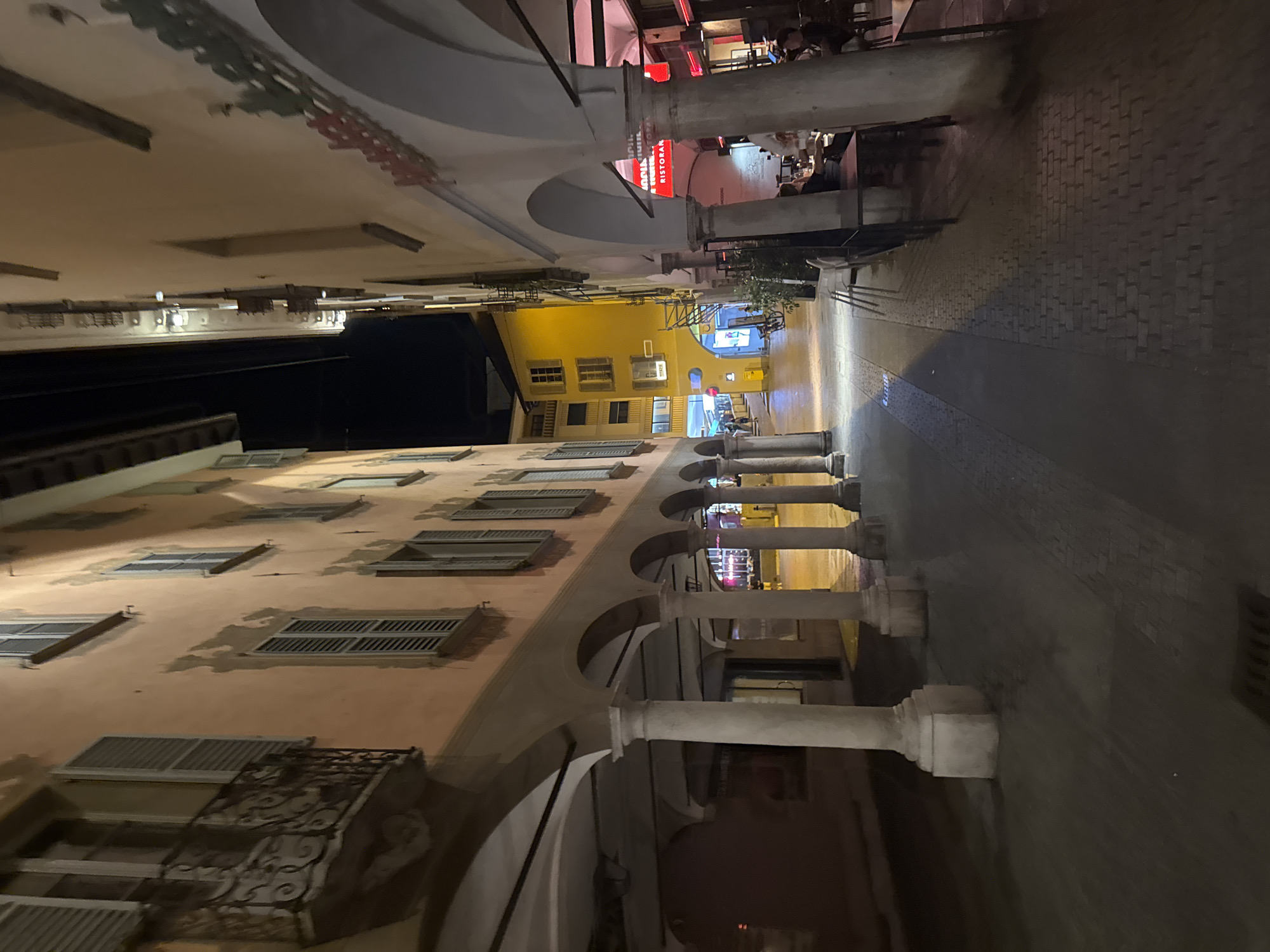 The image depicts a nighttime view of a narrow, arched porticoed street in Lugano, Switzerland. The architecture features a consistent style of rounded arches supported by thick, cylindrical columns, creating a covered walkway along the buildings. The buildings themselves are tall and slender, with windows that have shutters. The building on the right has a red neon sign that reads "RISTORANTE". There are tables and chairs set up under the portico, suggesting an outdoor seating area for a restaurant or cafe, with a few patrons visible in the background. The street is paved with cobblestones. The overall impression is a charming, historic European street scene at night, illuminated by artificial lighting.