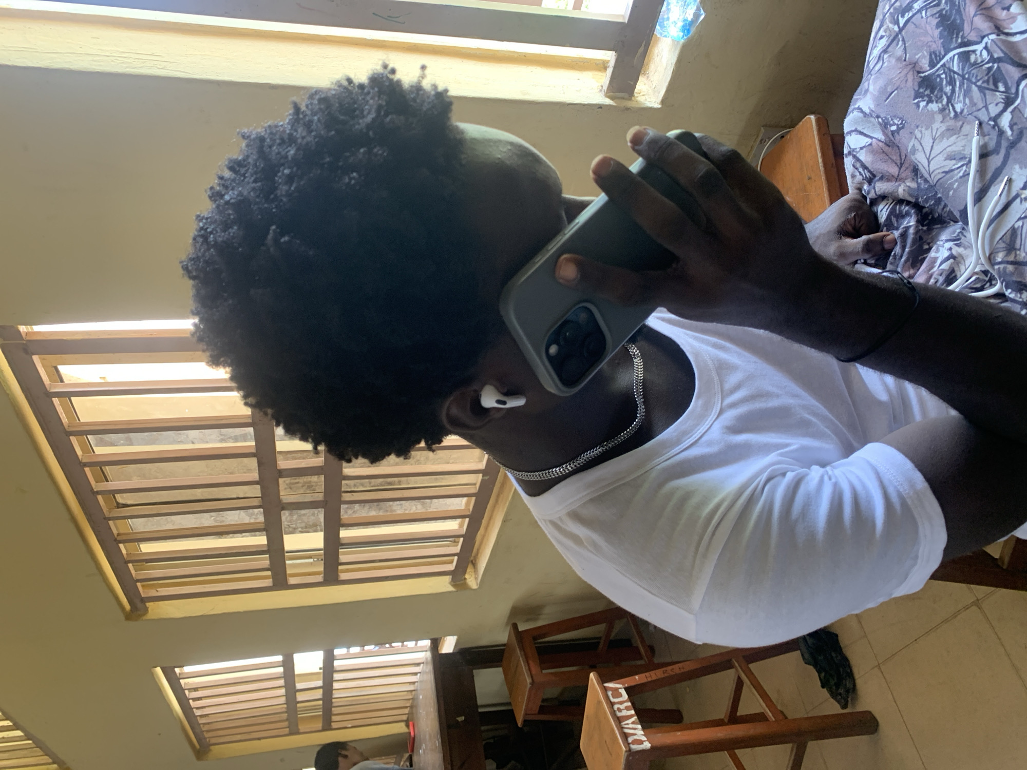 A person with dark, curly hair is holding a smartphone to their ear and speaking on it. They are wearing a white t-shirt and a silver chain. They also have white earbuds in their ears. The person is seated on a chair in a room with yellow walls and barred windows. There are wooden stools and a table in the background. The floor is tiled.