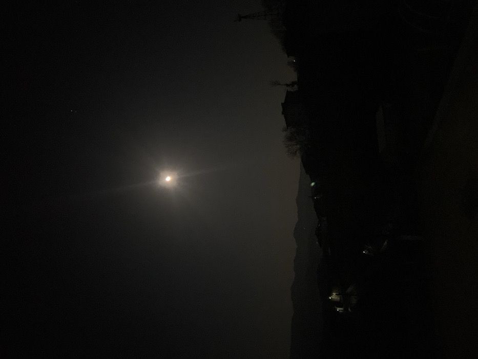 The image depicts a nighttime panoramic view of Sedlartsi, Bulgaria, under a clear, moonlit sky. The bright, nearly full moon, exhibiting a prominent lens flare, is positioned slightly above the central horizon line. Faint stars are visible in the dark sky, notably one above and to the right of the moon. The landscape consists of rolling hills or mountains forming a dark silhouette against the faintly illuminated sky in the background. In the mid-ground and foreground, the dark silhouettes of residential structures are visible, situated on elevated terrain. Several houses on the left exhibit visible exterior or interior lights, casting a soft glow. Bare trees are sparsely distributed among the structures and along the hillsides. On the far right, a distinct silhouette of a small wind turbine or similar structure stands against the sky on a higher elevation. No individuals, specific actions, or direct interactions are discernible within the scene. The overall environment is dark, with ambient illumination provided primarily by the moon.