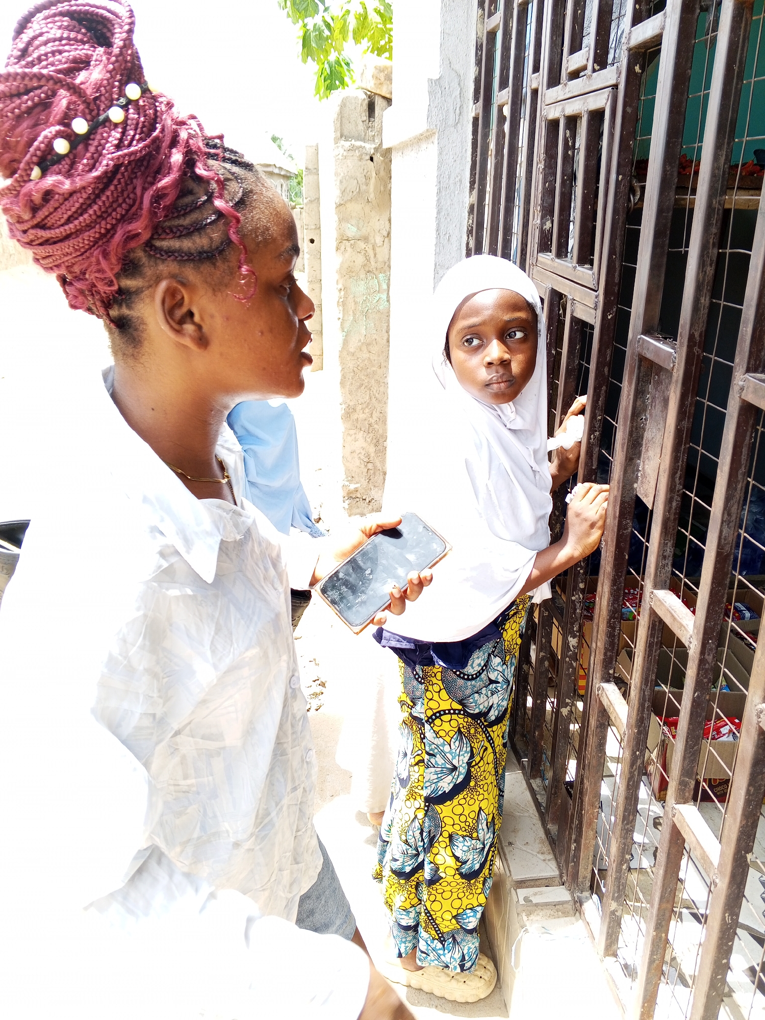 The image depicts a bright, sunny daytime scene in Adonkolo, Nigeria, featuring two main female subjects. On the left, an adult woman with elaborate reddish-brown braided hair styled in a high bun adorned with yellow and white beads stands, facing towards the right. She wears a light-colored, possibly white or light blue, collared button-down shirt and a delicate gold chain around her neck. She holds a dark smartphone in her right hand, and her mouth is slightly open, suggesting she might be speaking or about to speak.
On the right, a young girl is positioned behind a dark, rectangular-barred metal gate, peering out. She wears a white hijab and a vibrant, patterned dress or skirt in yellow, blue, black, and white. Her left hand holds a dark smartphone, while her right hand rests on the metal gate. She looks intently towards the left, her expression conveying curiosity or thoughtfulness. A third individual, wearing a light blue shirt, is partially visible in the background between the two main subjects.
The setting is outdoors, likely a street or an area adjacent to a building or shop. The bright sunlight indicates it is daytime, possibly mid-day or afternoon, creating strong highlights. Behind the gate, various items in cardboard boxes are visible, suggesting a commercial space. One box distinctly shows "CHOCO" and "PREMIUM" text, hinting at a chocolate product. The architecture includes light-colored, possibly concrete walls, and green foliage is visible at the top of the frame. The scene captures a moment of casual interaction or observation in a local community setting.