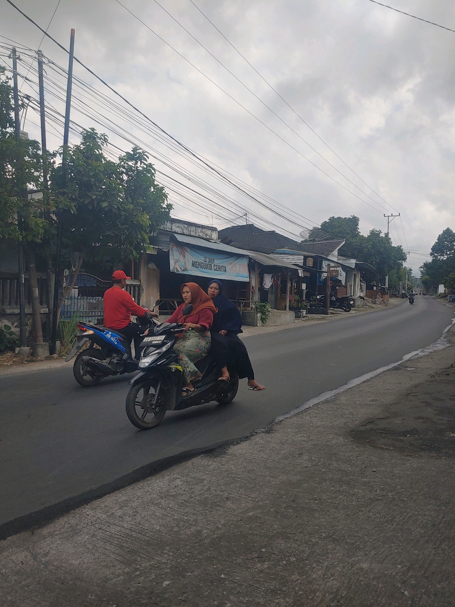 In Kembang Kerang, Indonesia, on a cloudy day, two motorcycles are traveling down a paved road lined with buildings and trees. A man wearing a red shirt and a red cap rides a blue motorcycle, while a woman in a red hijab and a patterned skirt, along with another woman in a dark hijab, ride a black motorcycle. They are all moving in the same direction. The buildings on the left appear to be shops or homes, some with signs. Overhead, numerous electrical wires crisscross the sky. The road surface is asphalt in the distance, transitioning to a rougher, gray concrete on the right side of the foreground.