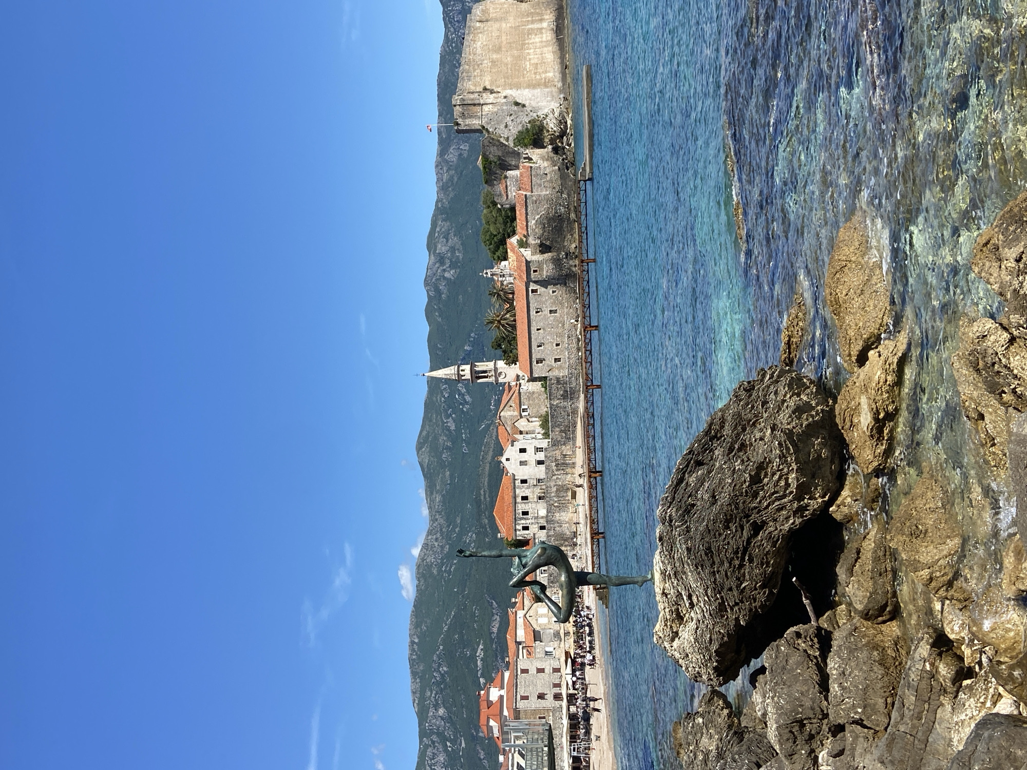 A vertical shot captures a bronze statue of a ballerina balancing on rocks with the city of Budva, Montenegro, in the background. The statue, rendered in a dark bronze color, is situated atop a rock formation at the edge of clear turquoise water. The old city of Budva features buildings with red-tiled roofs and a prominent church spire. Mountains rise behind the city. The sky is a clear, bright blue.
