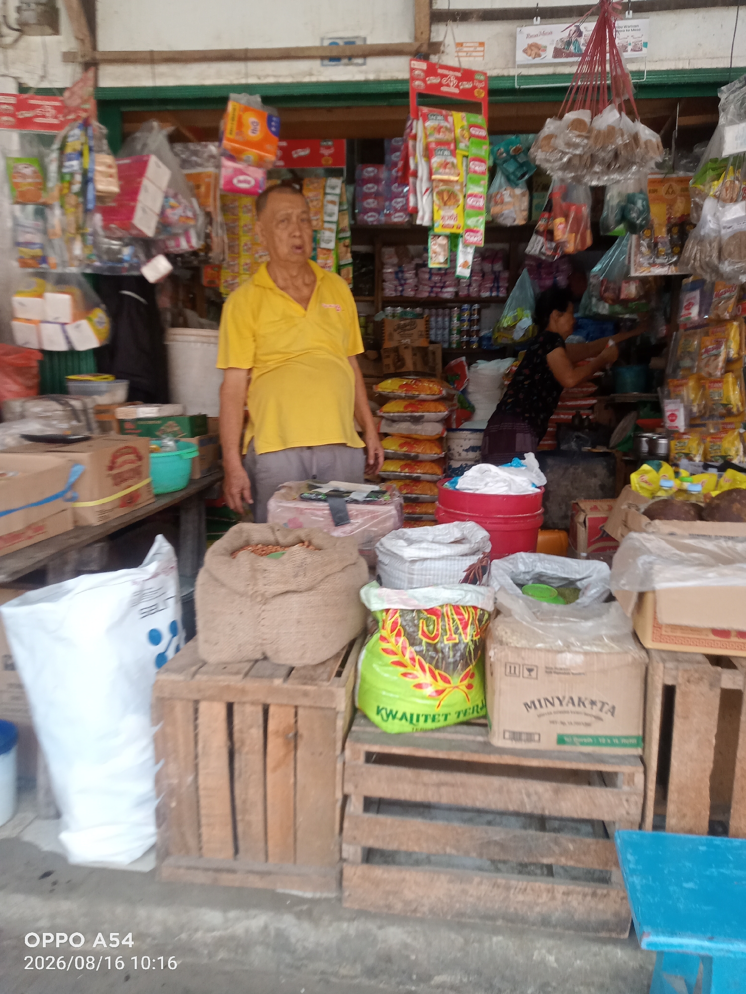 A middle-aged Asian man wearing a bright yellow t-shirt stands in front of a store. He is positioned in the center of the image and appears to be looking slightly to his left. He has short, graying hair and his face is somewhat obscured by shadow. His left hand is visible, resting at his side. Behind him, the store is filled with various products, including bags of rice, bottled goods, and packaged snacks displayed on shelves and in baskets. A woman wearing a dark floral shirt and a dark skirt is visible in the background on the right, appearing to be attending to the store's inventory. The store appears to be an outdoor market stall or a small, open-air shop, common in many parts of Indonesia, including Jambi. The floor is concrete. The lighting suggests it is daytime. The timestamp on the image indicates it was taken in Jambi, Indonesia, on August 16, 2026, at 10:16 AM.