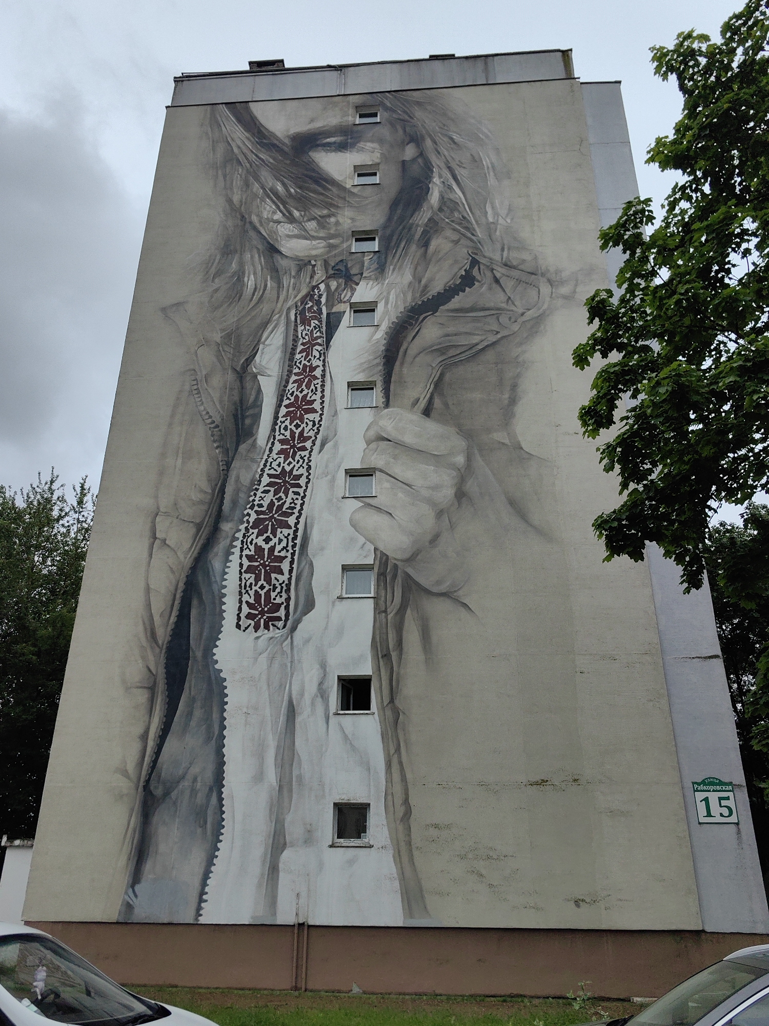 The image shows a large mural on the side of a tall apartment building in Minsk, Belarus. The mural depicts a grayscale portrait of a young woman with her hair blowing in the wind. She is wearing a white shirt with a red and black embroidered pattern around the neckline and chest, resembling traditional Slavic embroidery. Her left hand is shown near her face, touching her cheek or pulling at her collar. The building itself is light-colored, with several rows of windows visible on the muraled facade. To the right of the mural, a green street sign with white text reads "УЛИЦА РАБКОРОВСКАЯ 15," indicating the building is located at Rabkorovskaya Street, house number 15. A portion of a large green tree with dense foliage is visible in the upper right corner, partially obscuring the building. The sky is overcast and gray. The bottom of the image shows the lower part of the building, a brown base, a patch of green grass, and the hood and windshield of a white car.
