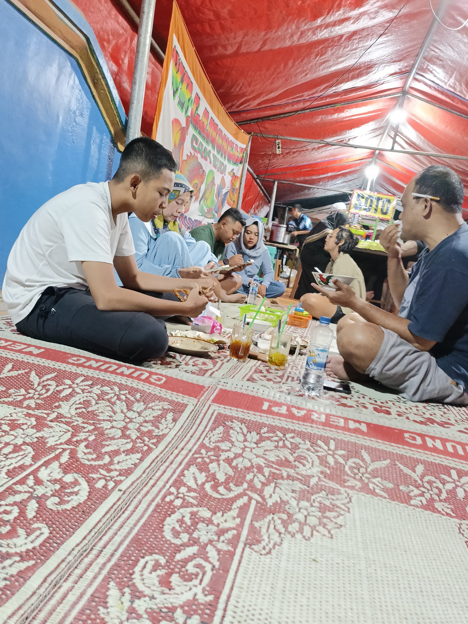 In Kebondalem Kidul, Indonesia, a group of six individuals are seated on a red and white woven mat under a red tarp canopy. The scene appears to be an outdoor food stall or gathering. The individuals are engaged in eating and socializing. A young man in a white t-shirt is on the left, holding food. A woman in a blue headscarf and blue top is next to him, also eating. Behind them, a man in a green shirt is looking at his phone. To the right, a man in a dark blue t-shirt and grey shorts is smoking a cigarette and holding a bowl with a picture of a rooster. Another individual is partially visible behind him. In the background, there are signs, food containers, and bottles of drinks. The overall environment is dimly lit, with artificial lights illuminating the area.