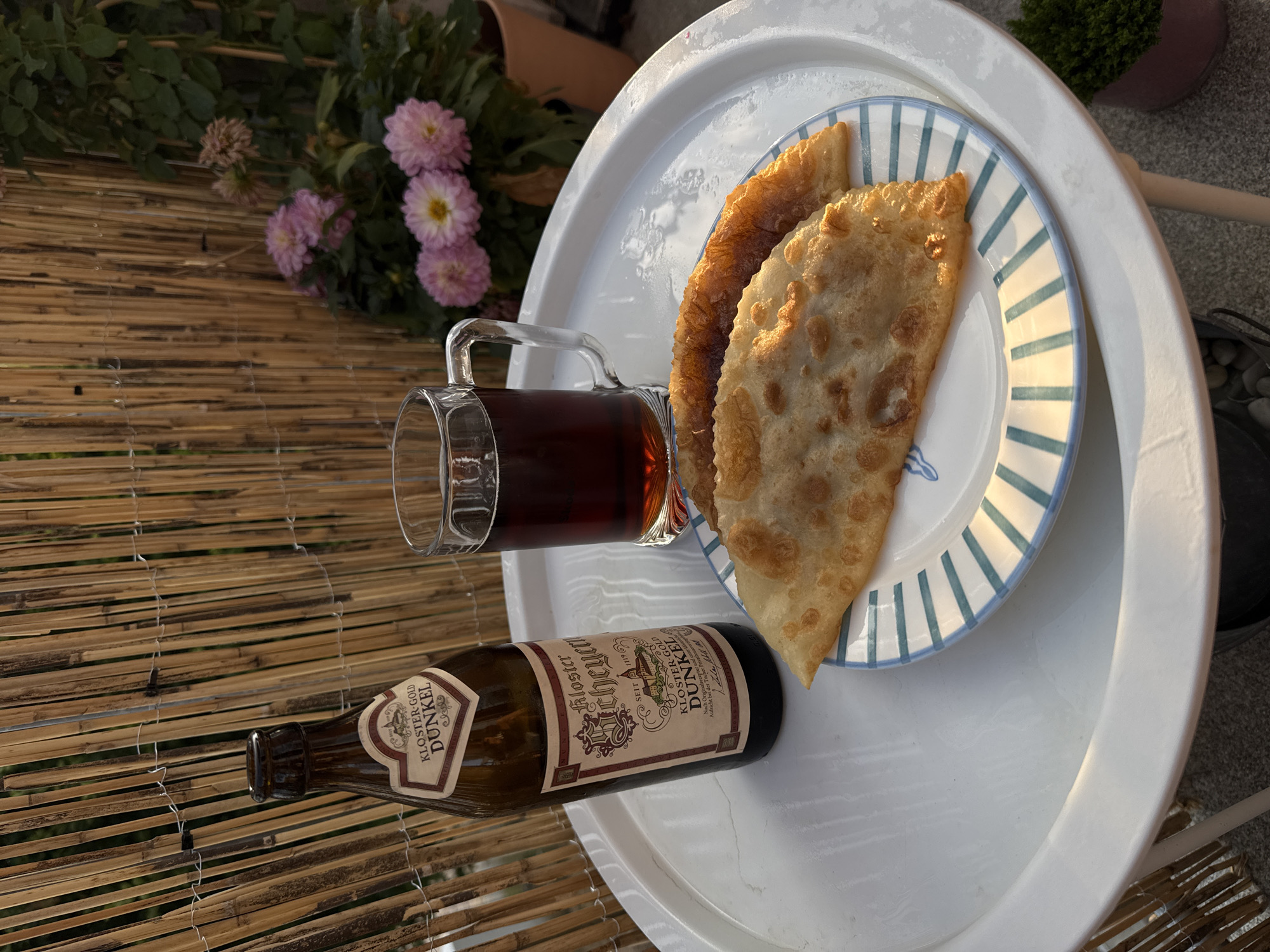 A bottle of Kloster-Gold Dunkel beer and two chebureki pastries are displayed on a white round tray. The tray is placed on a table with a woven bamboo fence in the background. Pink dahlias are visible behind the tray. The scene appears to be outdoors, possibly in a garden. There are no people visible in the image. The lighting suggests it is late afternoon or early evening.