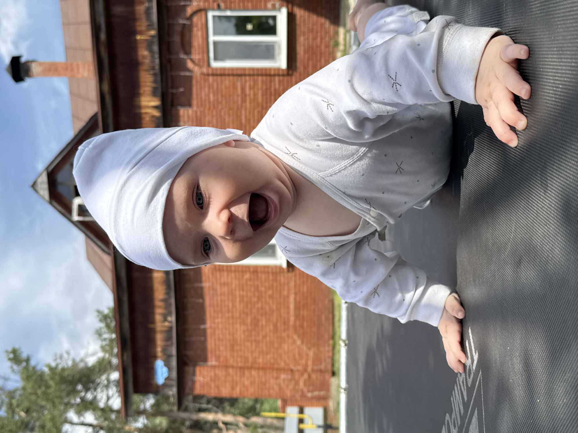 A baby wearing a white hat and onesie is lying on their stomach on a trampoline outdoors. The baby's mouth is open. The background includes a red brick building with a chimney, trees, and a blue sky with white clouds. The location is Rusalevka, Russia.