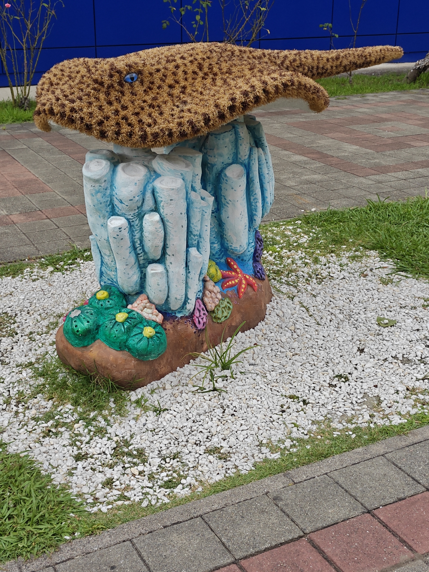 An outdoor, eye-level, medium shot captures a decorative sculpture of a stingray with a blue eye, sitting atop a base resembling coral or underwater rock formations. The stingray's body is covered in a brown, textured material, possibly coir or a similar natural fiber, with darker brown speckles. The base is painted in shades of blue and white, mimicking the appearance of underwater columns, and is adorned with green, yellow, and red sea-themed decorations including starfish and anemone-like shapes. The sculpture is situated on a bed of small white pebbles, with patches of green grass visible. In the background, there is a section of red and gray patterned pavement, a strip of green grass, and a blue wall. The overall setting appears to be an urban park or public space.