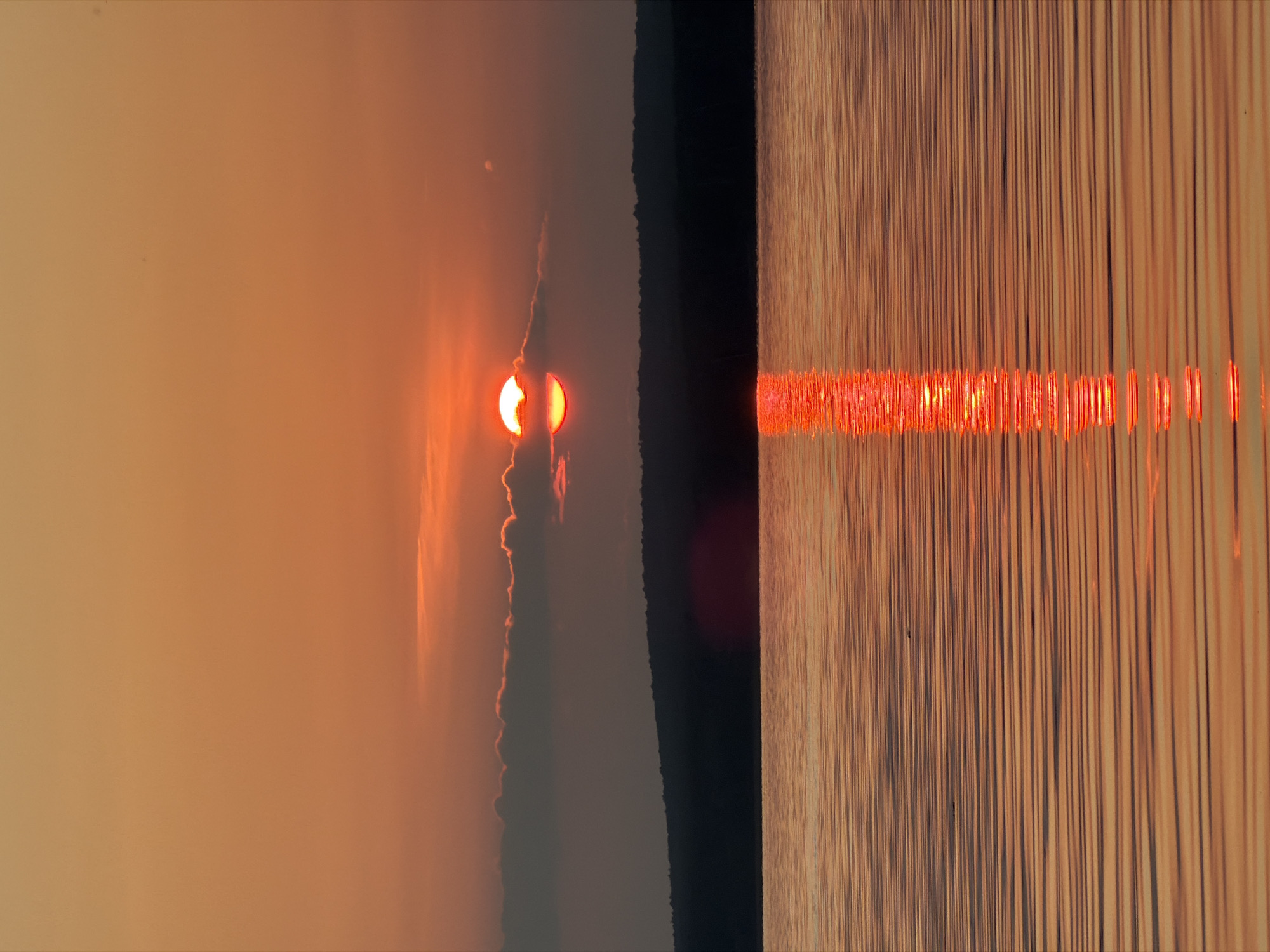 A sunset over a body of water with a dark, tree-covered hill in the background. The sun is a bright orange orb partially obscured by clouds, casting a reflection on the rippling water. The water is a warm orange-brown, reflecting the sky's colors. The scene is serene and tranquil, with no people or man-made structures visible. The image is a landscape photograph, capturing the natural beauty of the sunset. The location in the city of Chaikovsky, Russia, suggests this body of water is a river, lake, or reservoir within or near the city.