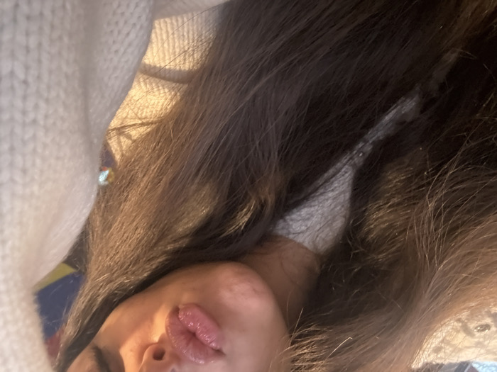 A close-up, angled view showcases a person lying down. Lush, dark brown hair dominates the frame, cascading around a face partially visible. The individual's lips are full and shiny. The person is wearing a chunky, off-white knitted sweater, adding a cozy texture to the left side of the image.