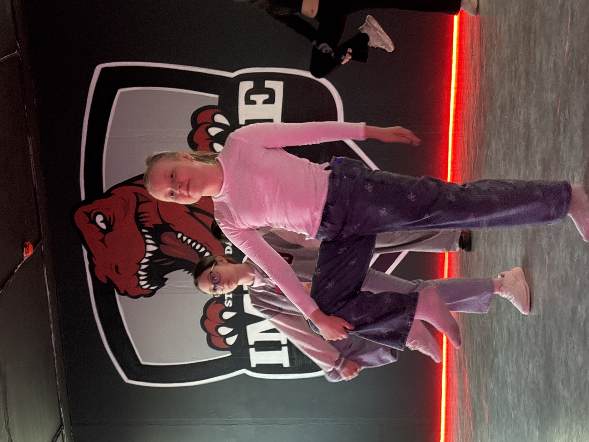 The image displays three young females performing stretching exercises within a dance studio in Pervomaysky, Russia. The studio wall is black with a large graphic featuring a red dinosaur head inside a shield, bearing the partial text "STUDIO DANCE IMAGINE". A bright red LED strip illuminates the base of the wall and the light gray floor. In the foreground, a blonde-haired female wearing a light pink long-sleeved top and dark blue patterned jeans is barefoot and holds her raised left shin with both hands. Behind her, a dark-haired female wearing glasses, a light top, a gray zippered hoodie, and light pants with white sneakers holds her raised right shin. Partially visible on the far right, a third individual in black athletic wear and light sneakers is also engaged in a leg-raising stretch.