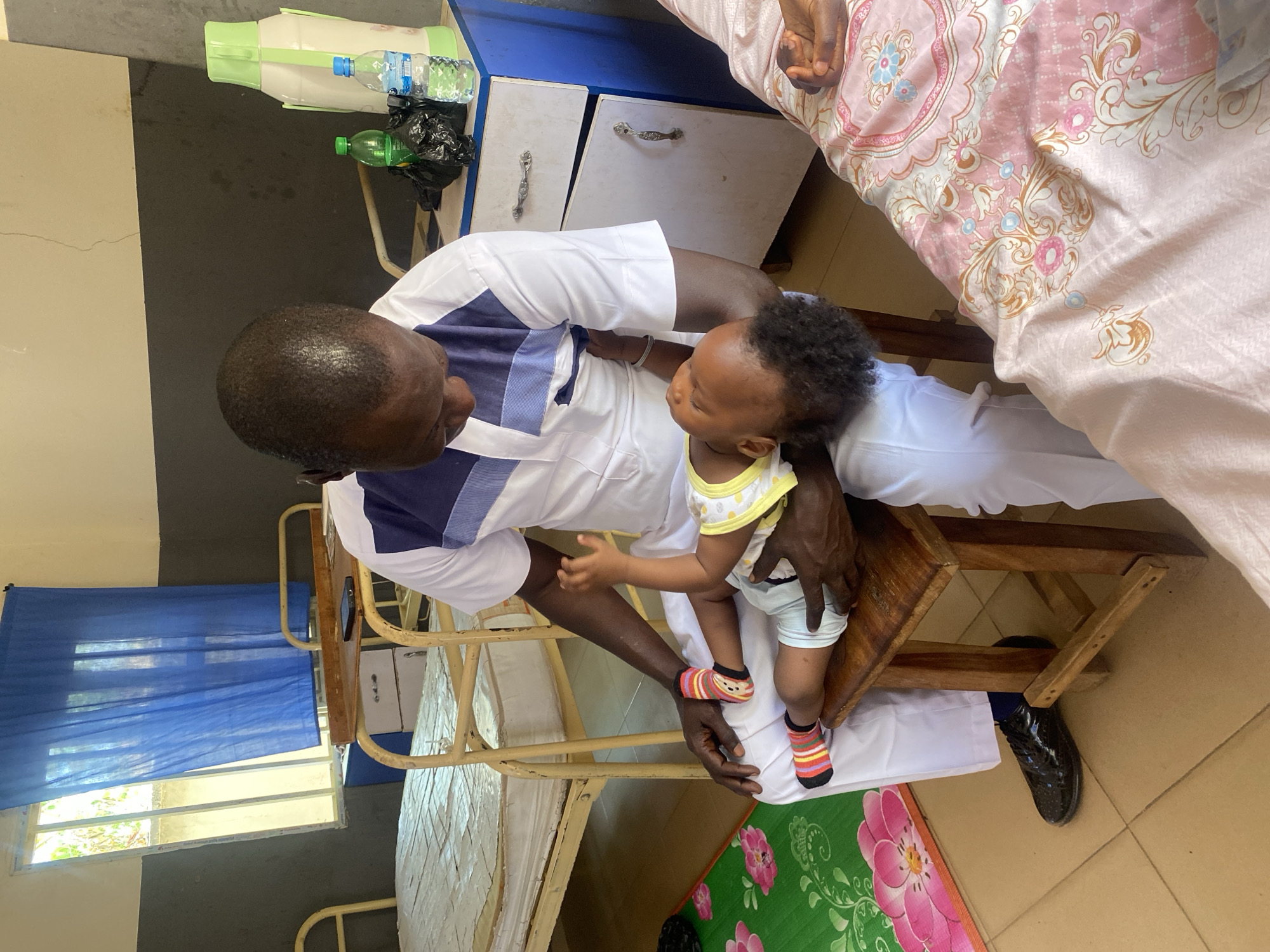 The image depicts an indoor scene, likely a hospital room, in Rido, Nigeria. An adult male with dark skin and short hair is seated, wearing a white uniform with a dark blue trim. He is holding a baby with dark hair, wearing a yellow and white onesie with striped socks. The baby is sitting on a wooden stool.
Behind the man, there is a hospital bed with a metal frame and a blue curtain partially covering the window. Next to the bed is a light blue cabinet with drawers. On top of the cabinet are bottles of water and a thermos.
The floor has tiled surfaces. A rug with a floral pattern is visible under the wooden stool. In the top right corner of the image, there is a bed covered with a pink floral bedspread.