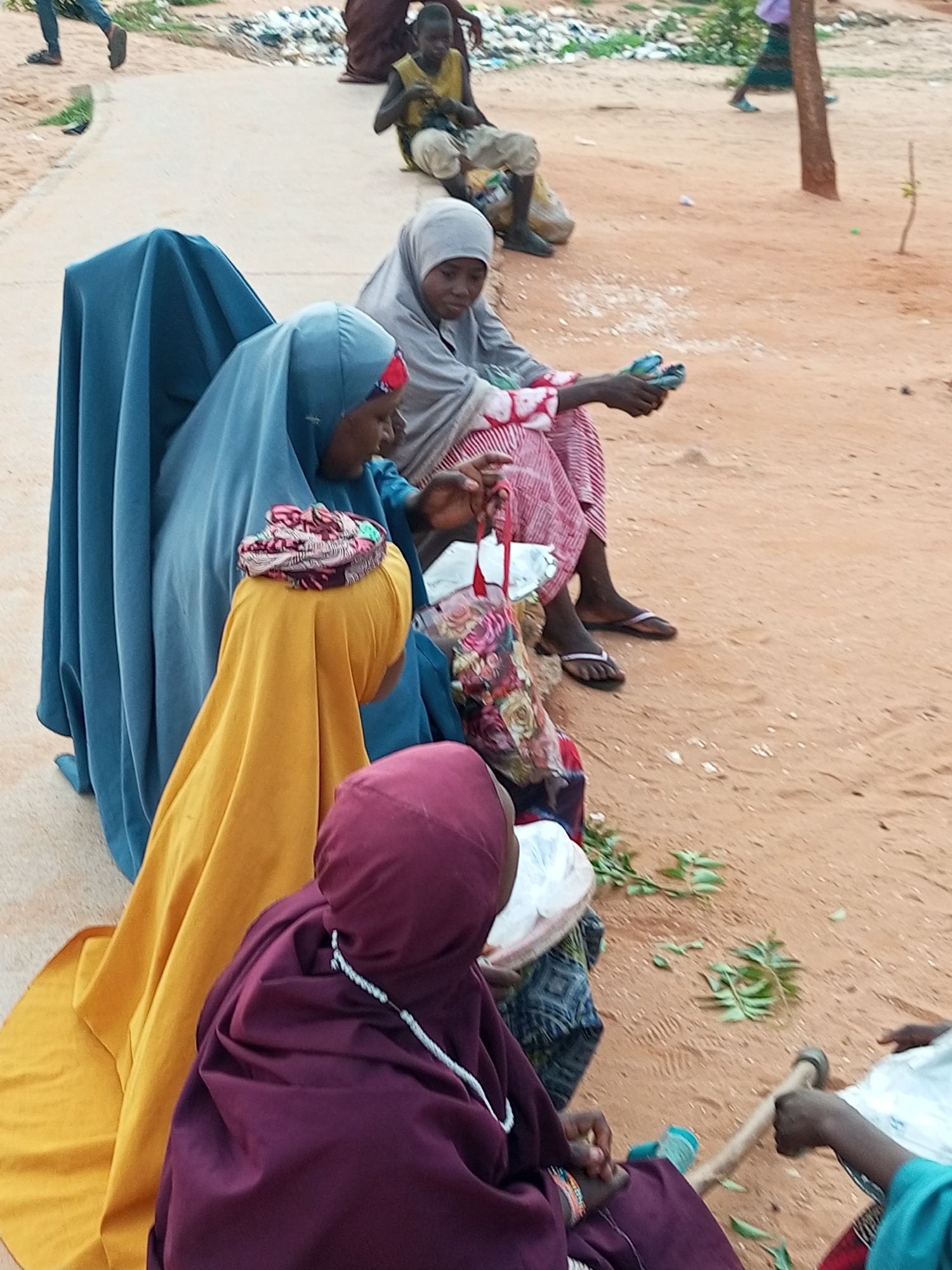 In Gombe, Nigeria, a group of people, primarily women and children, are gathered outdoors on a dirt ground with scattered debris and some vegetation. In the foreground, three women are seated. One, wearing a dark purple headscarf and garment, is looking down. To her left, a woman in a yellow hijab is also seated. Further left and slightly behind her, another woman wears a bright blue hijab and a blue garment. To the right of the yellow-clad woman, a woman with a grey hijab and a striped pink and white garment is holding a small object in her hands. Behind them, a child in a yellow vest sits on the ground, and another person in a dark brown garment is partially visible. The ground is a mix of sand and dirt, with a concrete path visible to the left. A wooden handle from a tool or implement lies on the ground near the lower right of the frame. The scene appears to be in an outdoor area that may be a market or a communal gathering space.