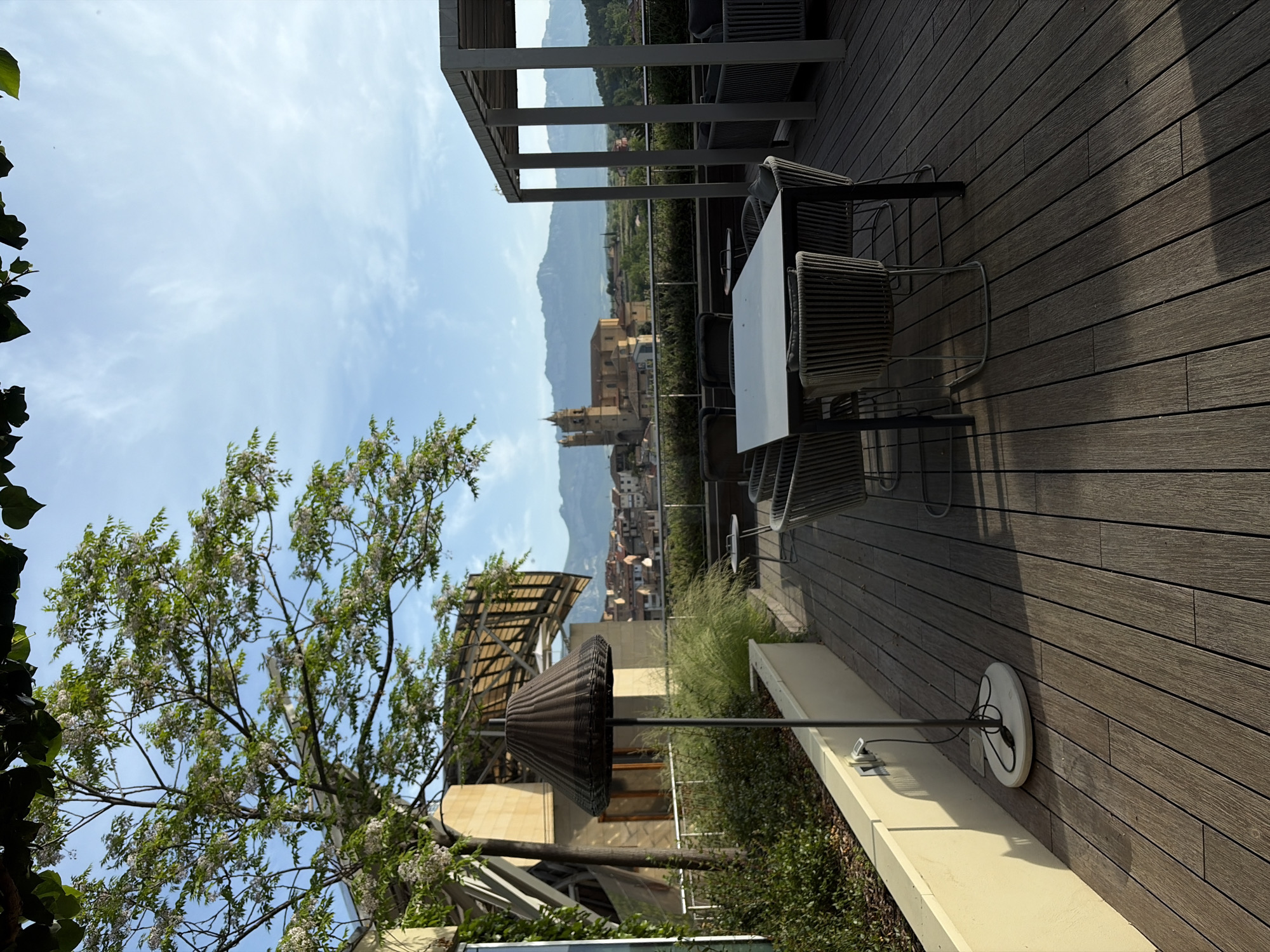 The image shows a rooftop setting, likely a restaurant or lounge, in Elciego, Spain, characterized by a wooden deck furnished with a table and chairs. Beyond the rooftop, the Spanish town is visible, with buildings and mountains in the background. In the foreground, a unique, woven shade structure can be seen. The sky is partly cloudy.