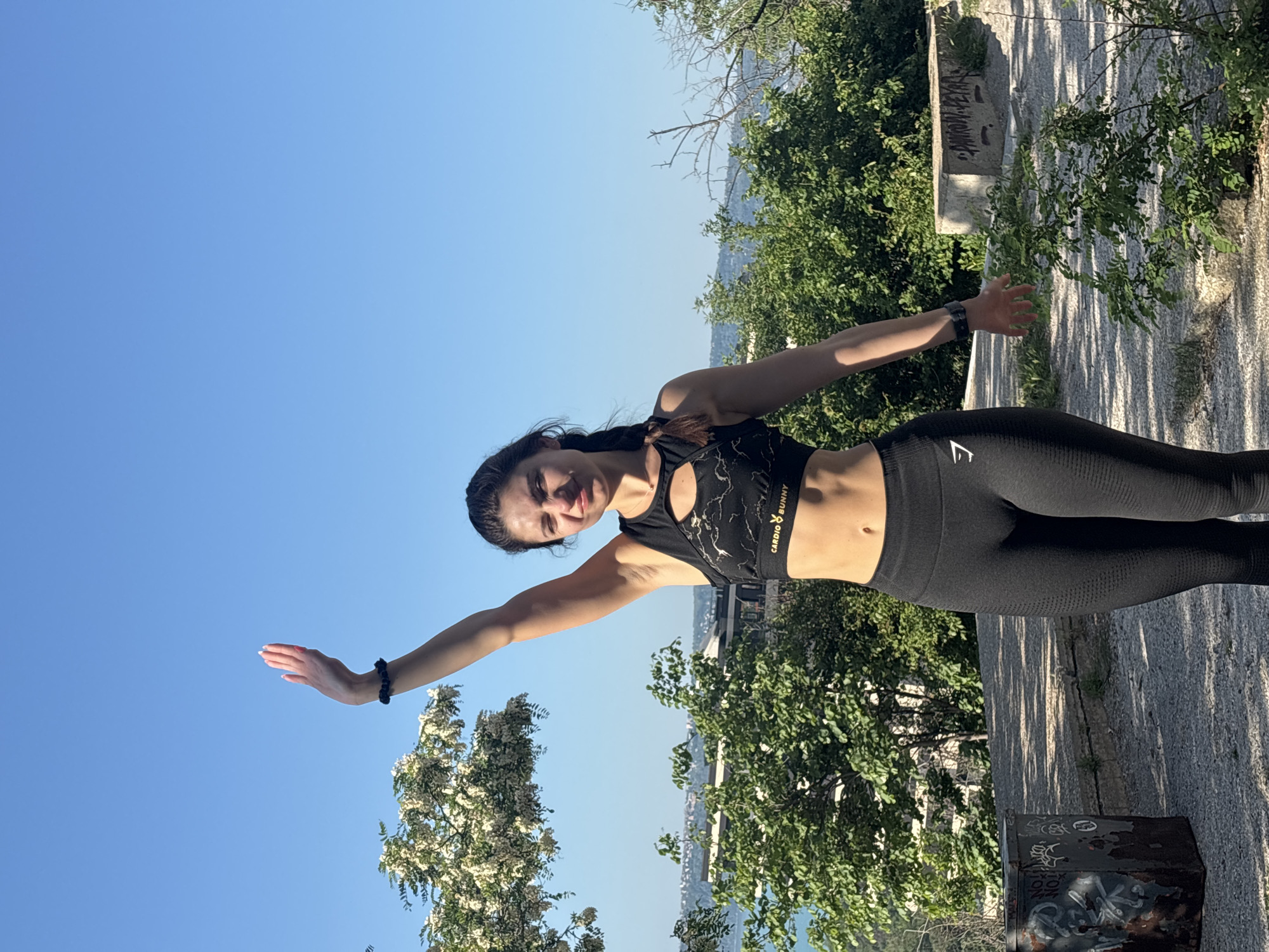 The image shows a woman standing outdoors on a concrete path, with trees and foliage surrounding her. She is wearing a black sports bra with a "Cardio Bunny" logo and black leggings with the Gymshark logo. Her arms are outstretched, with one hand pointing upwards towards the blue sky and the other pointing towards a stone wall. There is a cityscape visible in the background, and the location appears to be Varna, Bulgaria. A metal box with graffiti is also present on the concrete path. The lighting suggests it is daytime with clear skies.