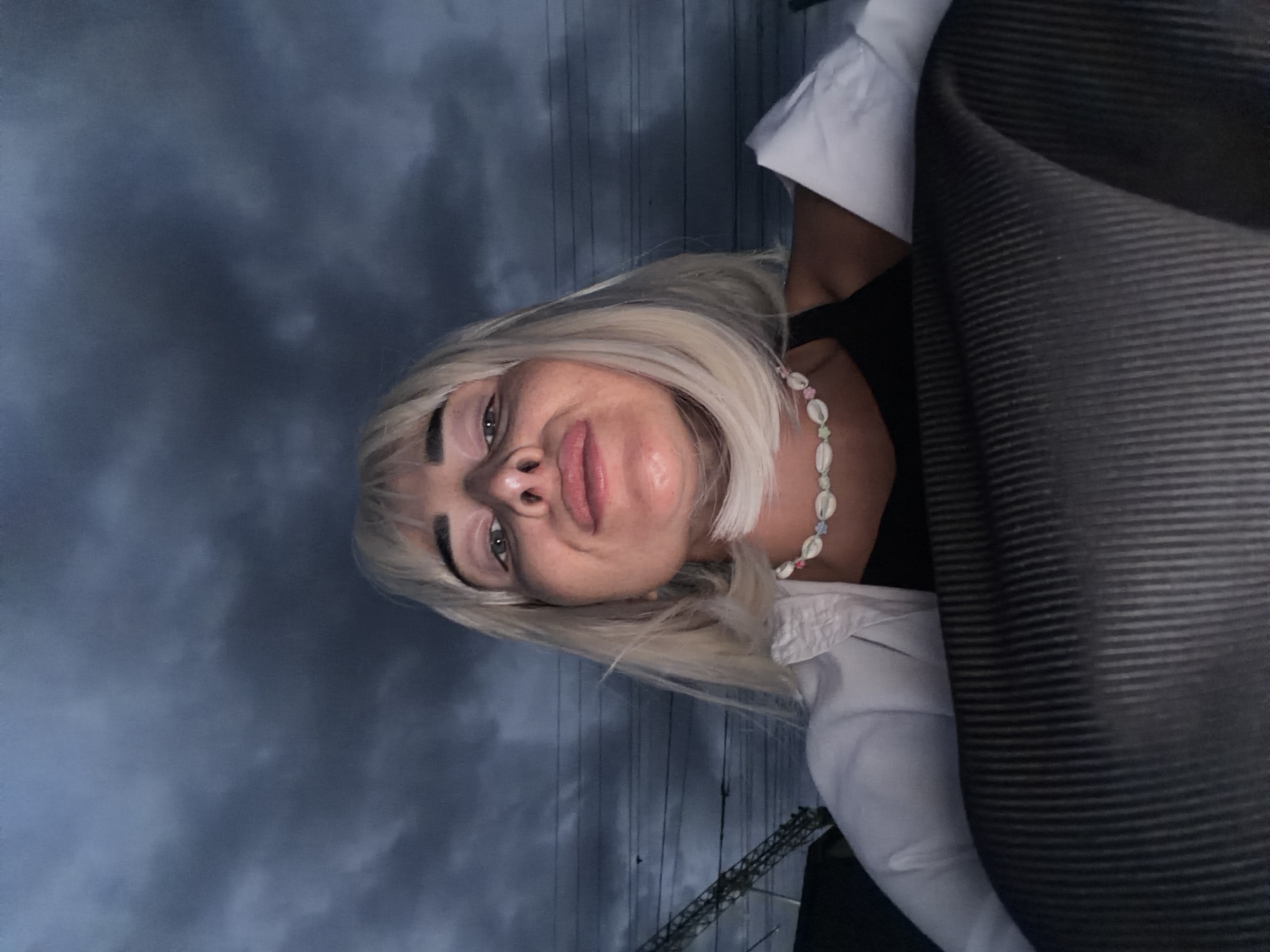 A woman with blonde hair and a necklace is shown from the chest up, leaning forward to take a selfie. She is wearing a white shirt over a black top. The background consists of a dark, cloudy sky with power lines and a structure that resembles a crane or tower. This scene is set in Sochi, Russia, as indicated by the presence of the power lines which are characteristic of the city's infrastructure. The image is taken at dusk, as suggested by the dim lighting and the dark clouds. The woman's pose and facial expression suggest she is looking at the camera, and the overall mood is somewhat dramatic due to the stormy sky.