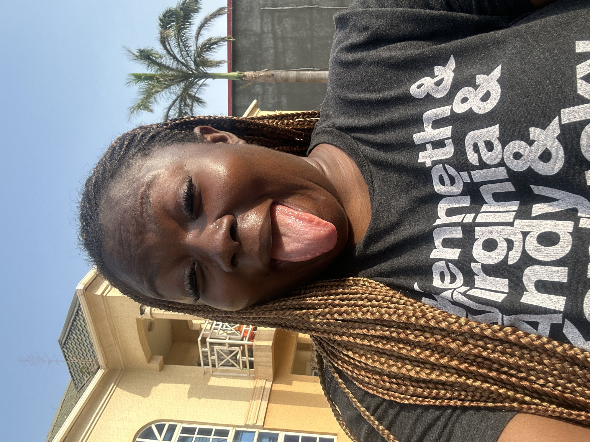 In a selfie taken outdoors under a clear blue sky, a Black woman with braided hair sticks her tongue out at the camera. She is wearing a dark gray t-shirt with white text that reads "Kenneth & Virginia & Andy &". The woman's eyes are closed, and she is wearing false eyelashes. Behind her, to the left, is a cream-colored building with arched windows and a balcony. To the right of the building, a palm tree stands tall. The ground is paved, and a concrete wall is visible behind the palm tree. The overall lighting suggests it is daytime, possibly late afternoon due to the warm light. The image is taken from a head-and-shoulders perspective.