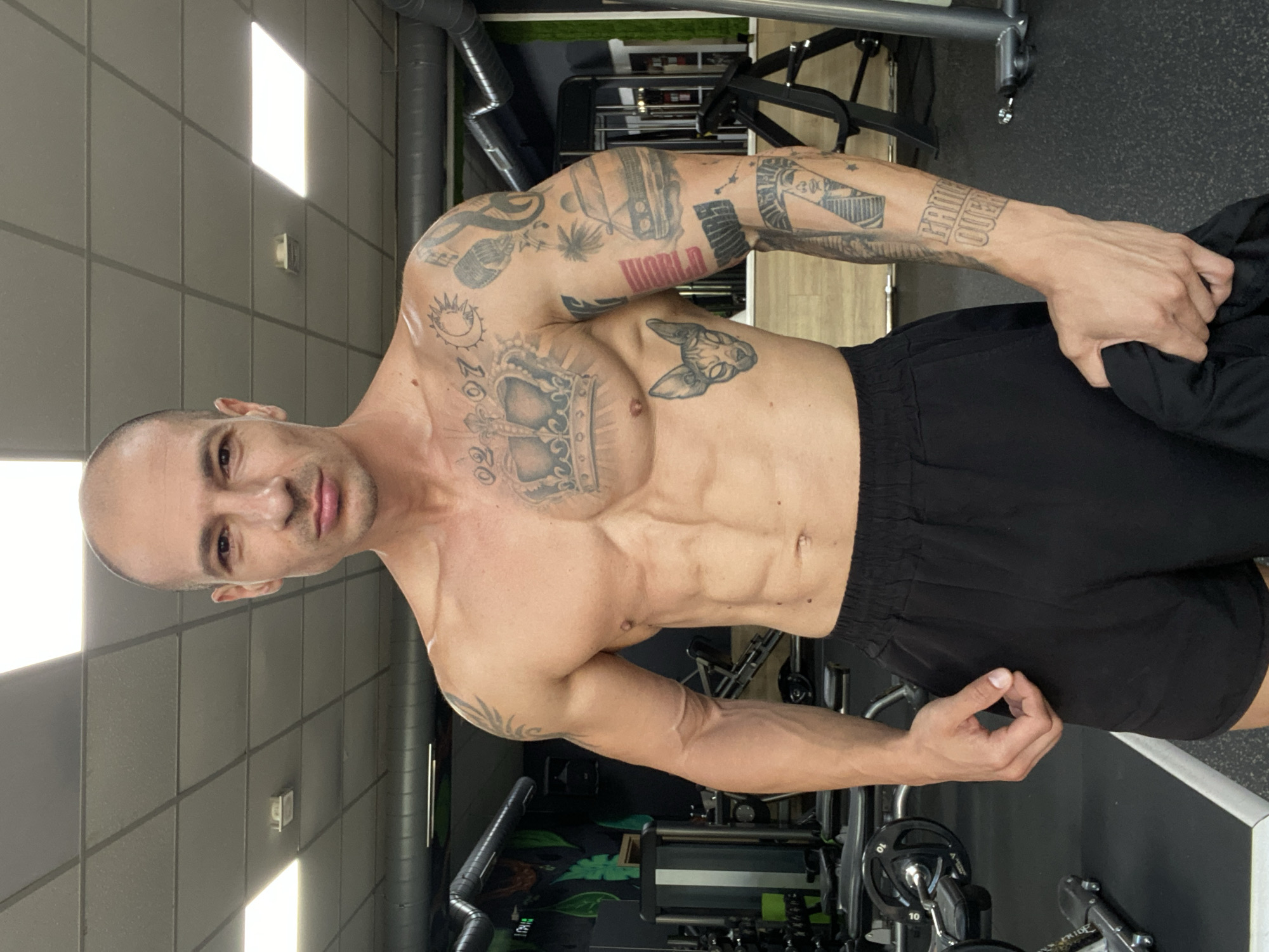 A fair-skinned, bald man with a muscular physique and numerous tattoos is standing in what appears to be a gym. He is wearing black shorts and is shirtless. His tattoos include a crown on his chest, a cat's face on his torso, and various images on his arm, including a car, a pharaoh, and the words "GAME OVER." The gym environment is visible in the background, with weightlifting equipment and fluorescent lighting. The image does not provide any specific information or visual cues to indicate the location as Pleven, Bulgaria.