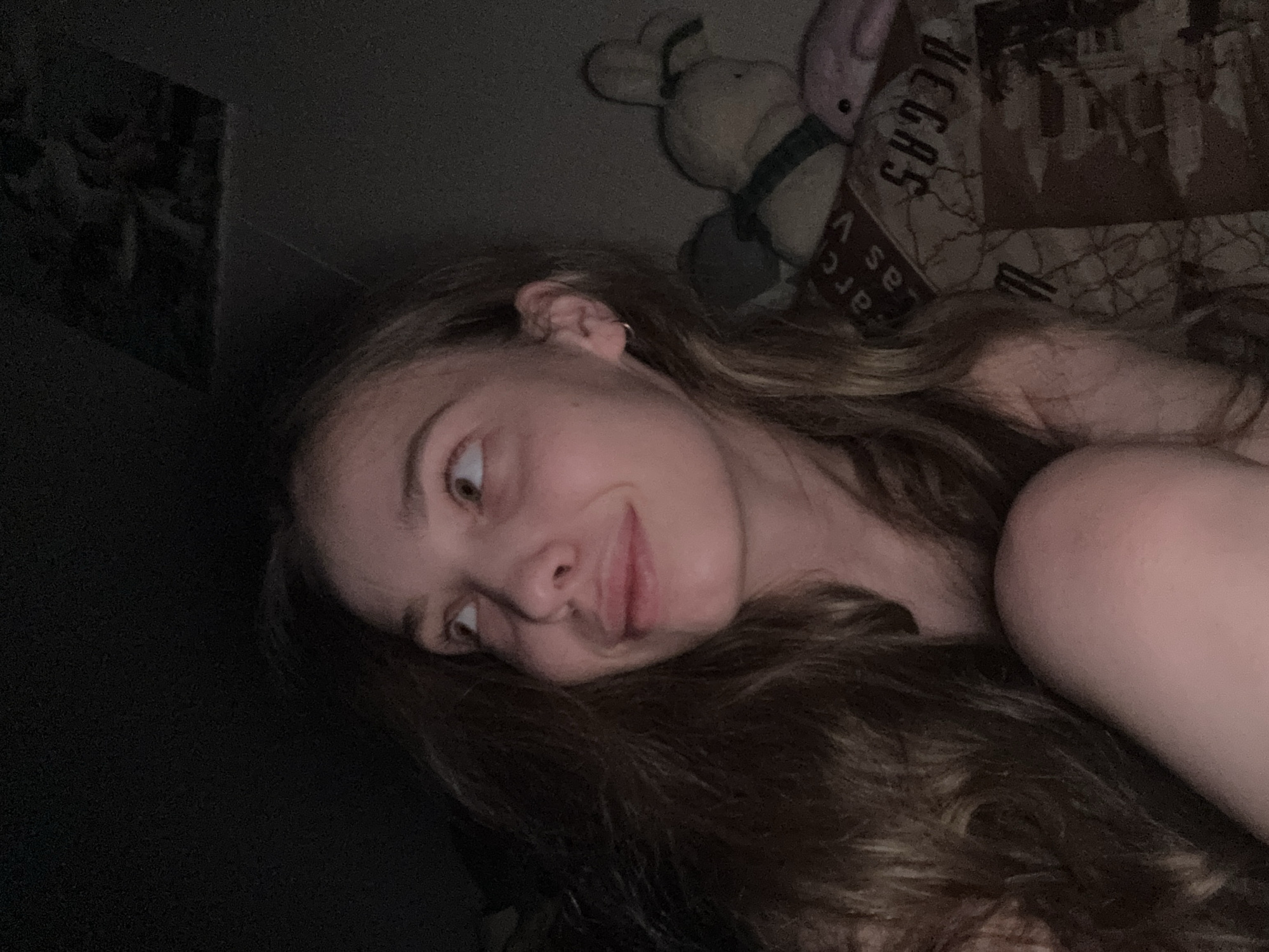 An individual, appearing female, with long, wavy light brown hair, is depicted in a dimly lit indoor setting. Her head is angled slightly to her left, and her eyes are directed upwards and to the right, accompanied by a subtle smile or smirk. A small silver hoop earring is visible in her right ear. Her bare right shoulder and a portion of her bent right knee are visible in the foreground.
The background to her right features patterned bedding or a blanket, with discernible, mirrored text including "arc as V" and "VEGAS," alongside faint graphic representations of urban structures, suggesting a Las Vegas theme. Resting on the bedding behind her are two plush toys: a light-colored rabbit with a dark green element around its neck, and a smaller, less distinct pink or purple plush. The upper left and top portions of the image are largely dark and indistinct, with faint traces of objects or patterns on a wall or ceiling. The overall lighting is low, casting soft shadows and indicating an indoor environment, likely a bedroom. The specific geographical location, including any city like Chaikovsky, Russia, cannot be determined from the visual information provided within the image.