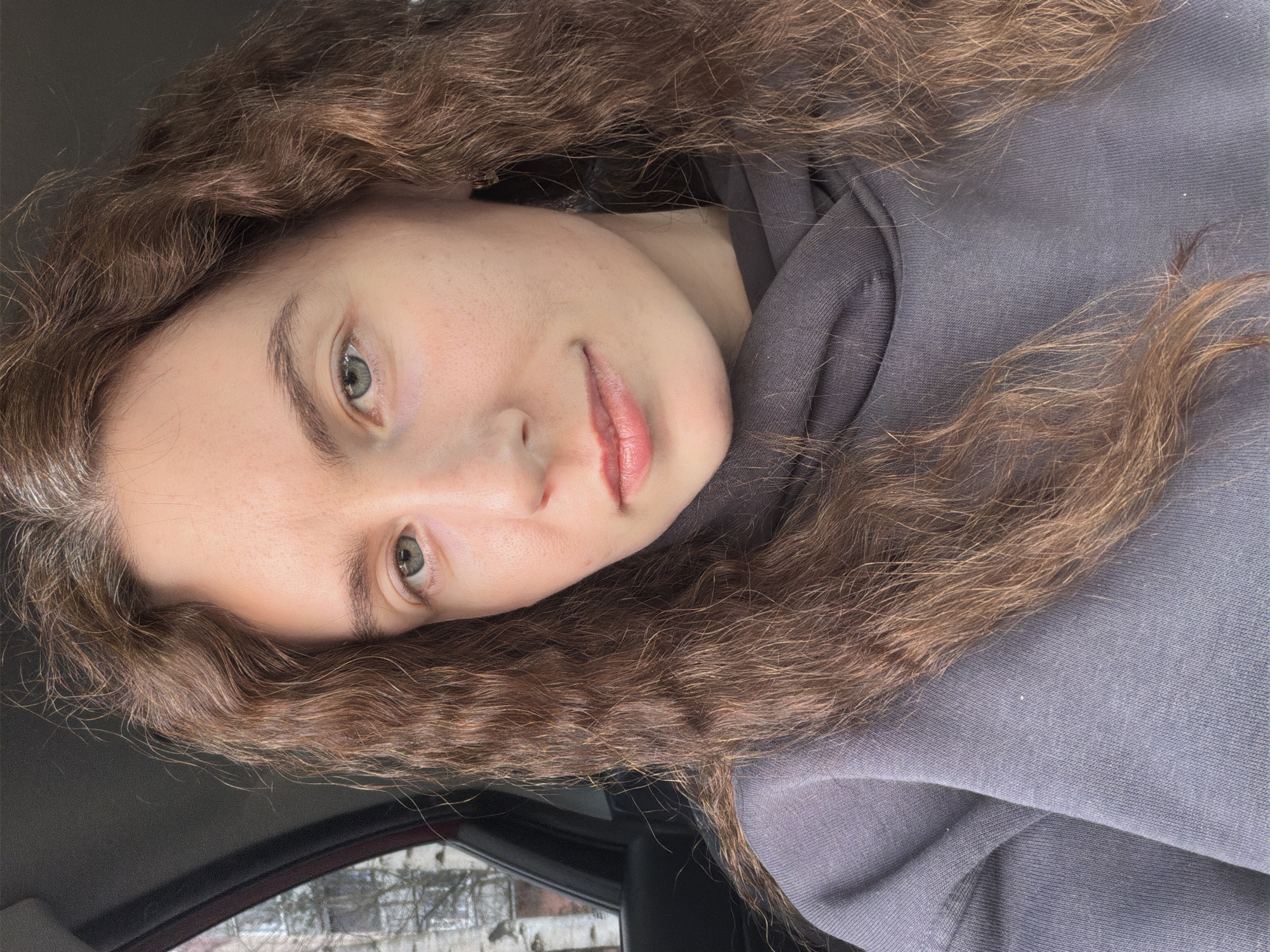 A close-up portrait of a young woman with light skin, light eyes, and wavy brown hair. She is wearing a gray hoodie. The background visible through the car window appears to be an urban environment, potentially a residential area, with trees and buildings visible, which suggests this photograph was taken in Vsevolozhsk, Russia. The interior of the car is visible, with parts of the dashboard and door visible in the lower left. The lighting is soft and diffused.