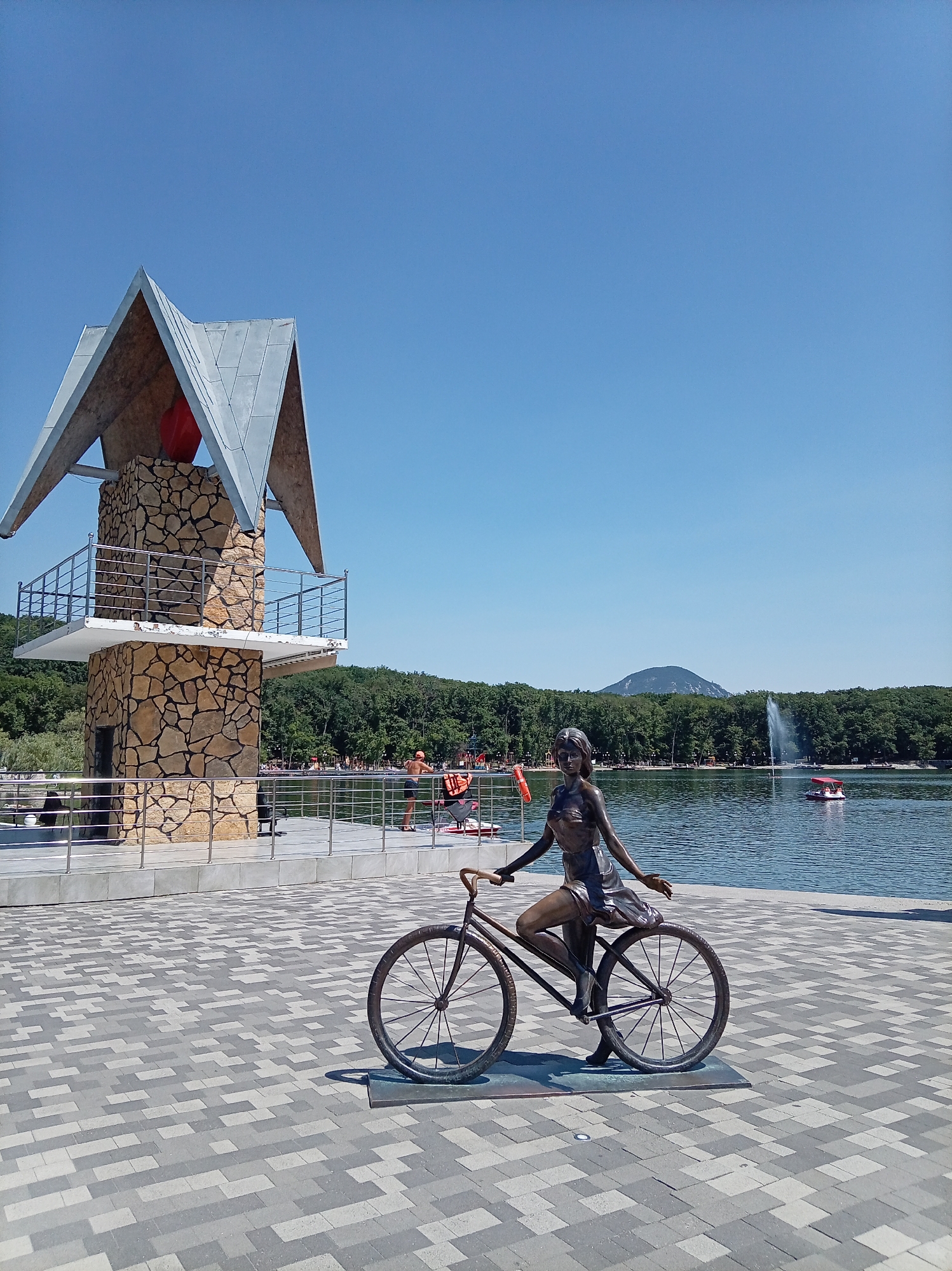 In the city of Zheleznovodsk, Russia, a bronze sculpture of a young woman on a bicycle is positioned on a paved walkway near a lake. The sculpture is in the foreground, with the woman appearing to be mid-pedal. Behind the sculpture, a stone tower with a distinctive angular roof structure stands next to a railing that overlooks the lake. The lake is calm with a few small boats and a water fountain in the distance. A line of trees borders the far side of the lake, and a mountain is visible on the horizon under a clear blue sky. Several people are visible on the walkway and near the lake, some wearing orange life vests, suggesting a recreational area.