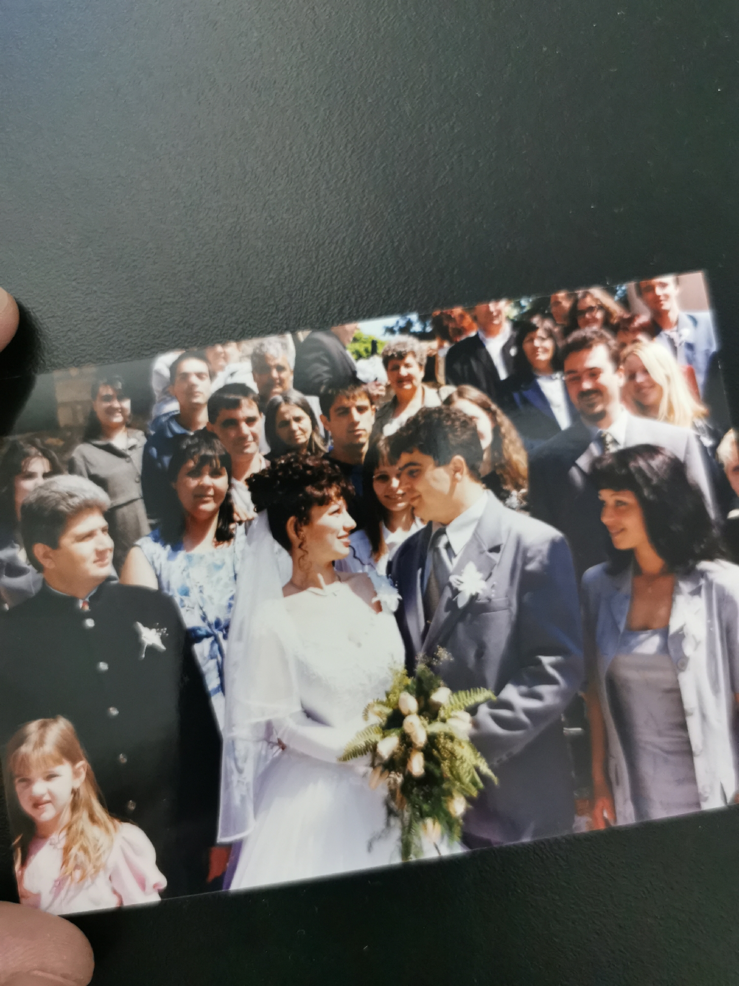 A photograph shows a group of people, including a bride and groom, posing for a wedding photo. The groom is in a light-colored suit with a flower on his lapel, and the bride is in a white wedding dress with a veil. She's holding a bouquet of flowers. Other people are in the background, dressed in various formal and semi-formal attire. A younger girl in a pink dress is at the bottom left corner of the photo. There is no visible evidence in the photograph to confirm the location is Varna, Bulgaria.