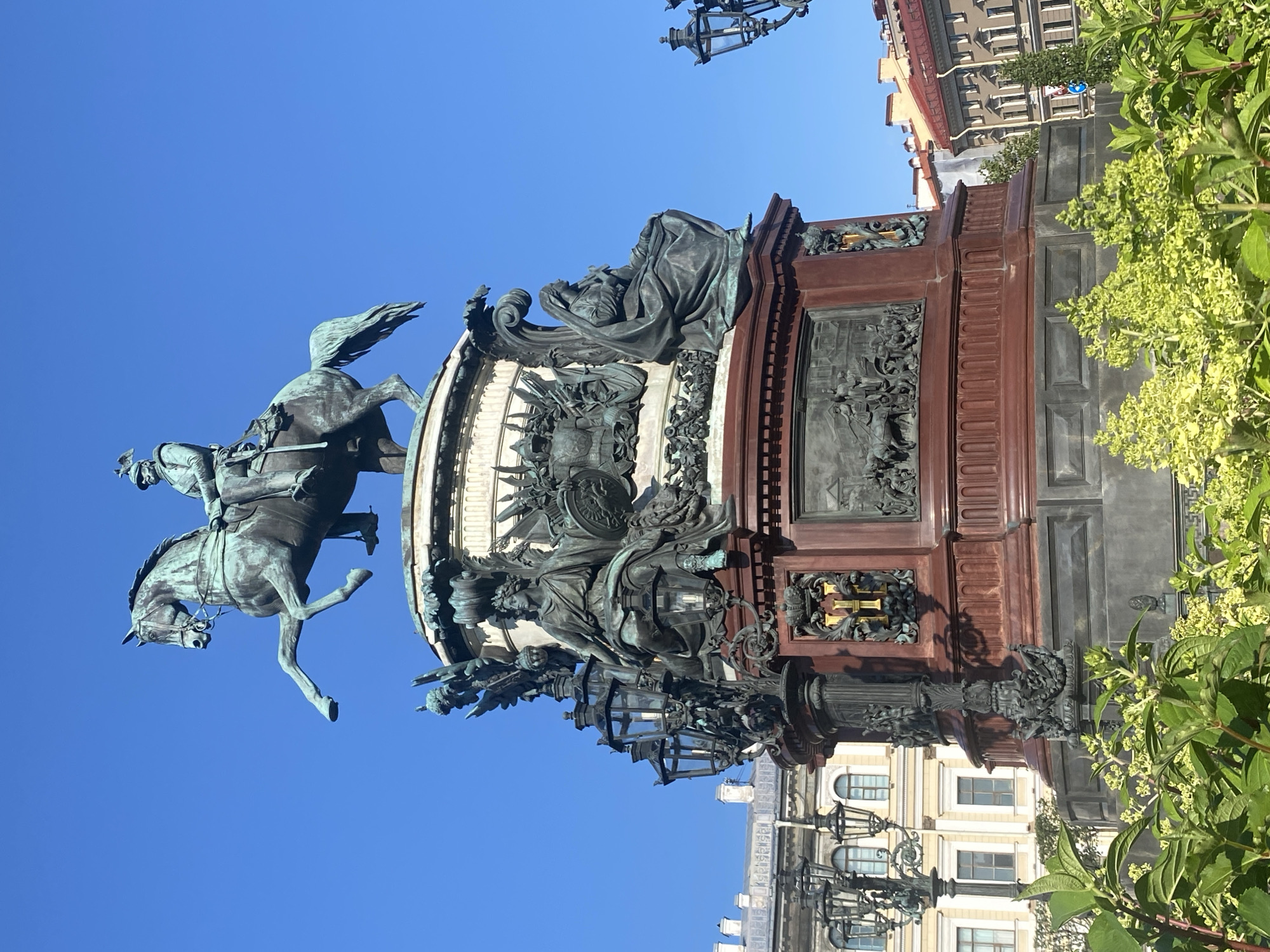 The image depicts the Bronze Horseman, a statue of Peter the Great on horseback, located in Senate Square, Saint Petersburg, Russia. The statue is made of bronze and stands on a tall pedestal. Peter the Great is depicted wearing a military uniform and a laurel wreath, with his horse rearing up on its hind legs. The pedestal is adorned with bas-reliefs depicting scenes from Peter the Great's reign.
Surrounding the statue are buildings with classical architecture, typical of Saint Petersburg. The sky is a clear, bright blue, indicating a sunny day. In the foreground, lush green foliage and flowering plants partially obscure the base of the monument. The overall scene captures the grandeur and historical significance of this iconic landmark.
