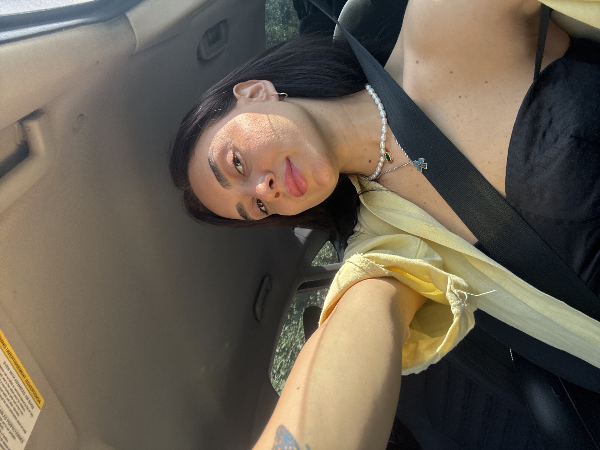 In a medium shot, a woman with dark hair and fair skin takes a selfie while seated in the driver's seat of a car. She is wearing a yellow shirt and a black top, along with a pearl necklace and silver earrings. A black seatbelt is fastened across her chest. The interior of the car is visible, with the dashboard and a portion of the passenger seat in the background. Sunlight streams through the car's windows, casting shadows on her face and body. The scene appears to be set during the day, possibly in an urban environment, as trees and foliage are visible through the car windows, suggesting a location in Tbilisi, Georgia.
