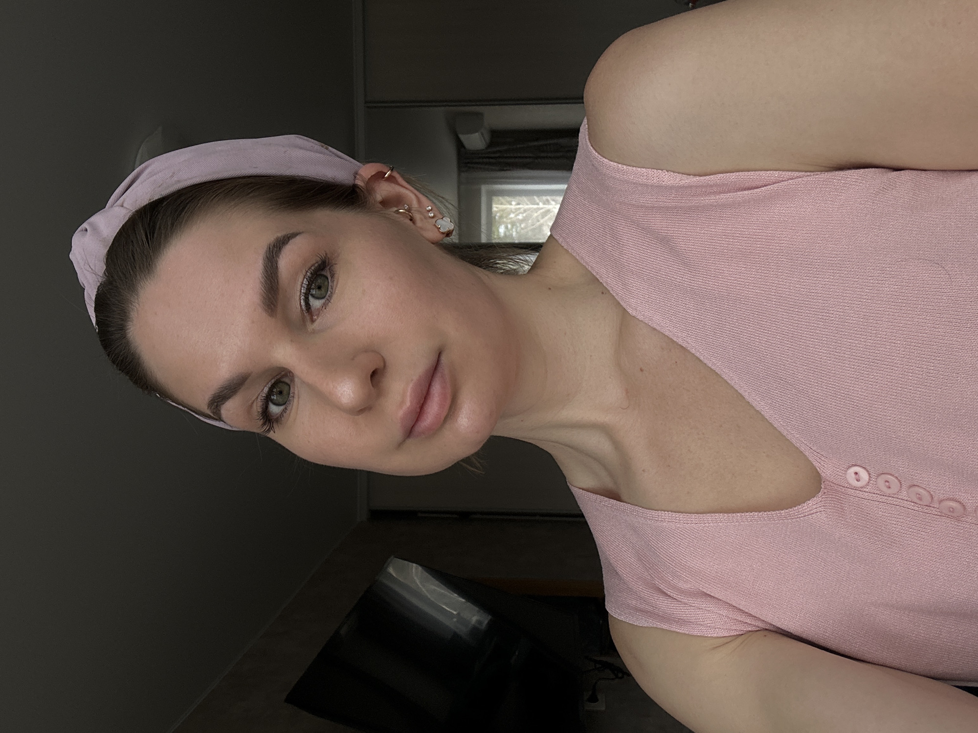 A woman with light skin and brown hair pulled back by a light pink headband poses for a close-up selfie. She is wearing a light pink top with small buttons down the front. She has several earrings in her left ear. The background is dark, with a television screen partially visible and a window with a view of trees and other buildings. The location is not indicated in the image.