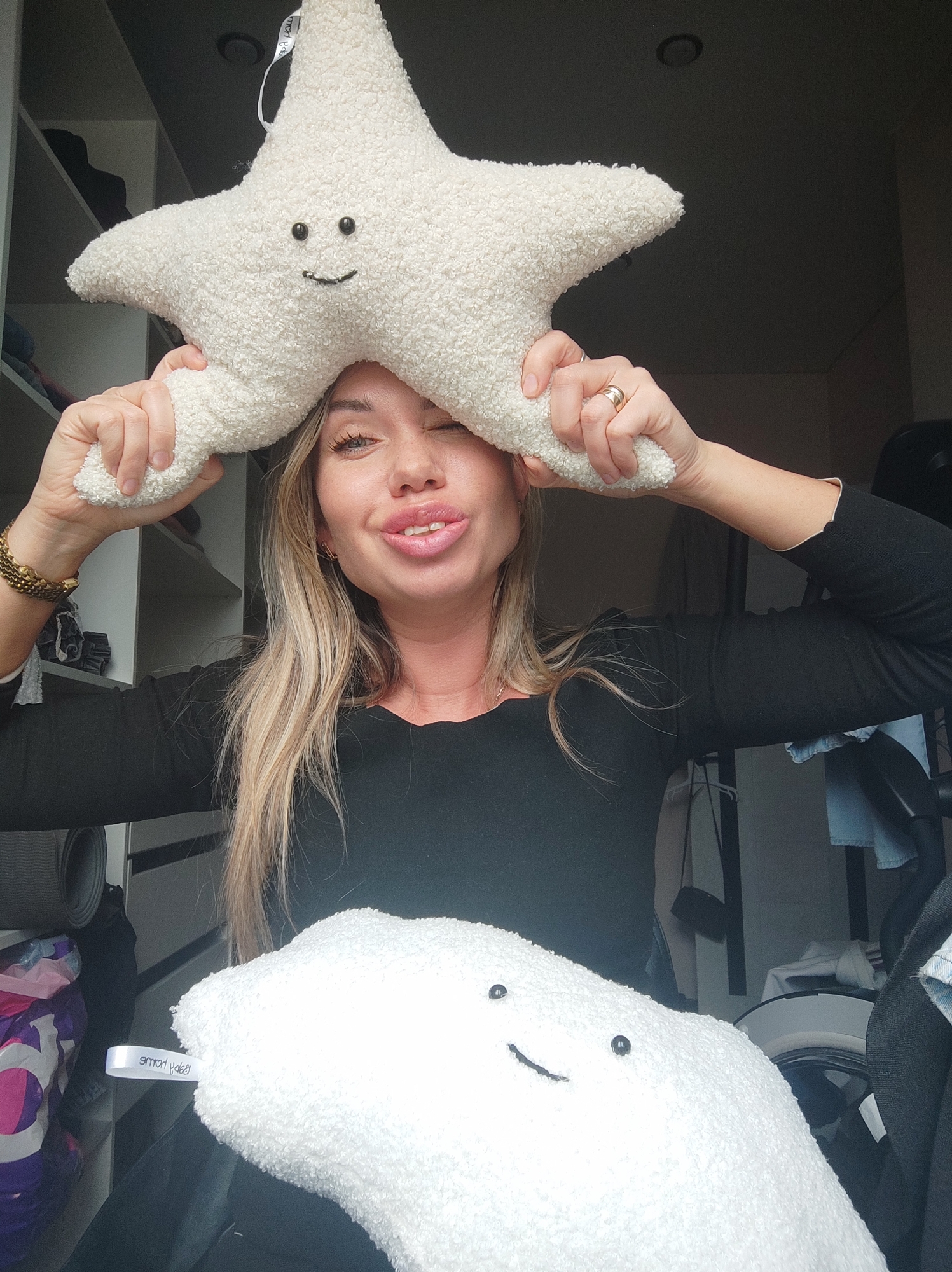A woman is holding a white, star-shaped pillow with a smiley face above her head, while also holding a white, cloud-shaped pillow with a smiley face below her chest. She is winking and making a kissing face. She is wearing a black long-sleeved shirt and a gold watch on her left wrist. Behind her are shelves, clothing, and other items, suggesting a closet or bedroom setting. It is not possible to determine if the location is in Votkinsk, Russia.