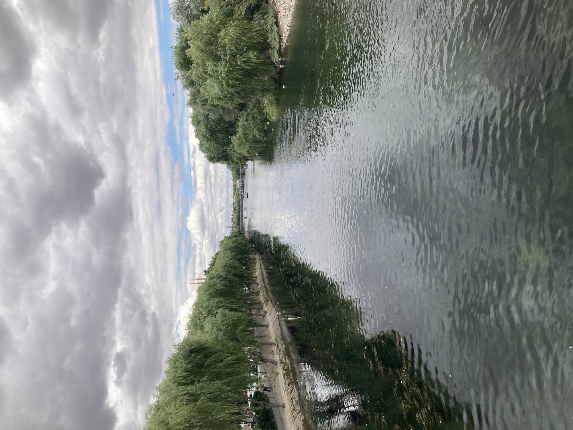 A wide, calm river flows through the city center of Avanos, Turkey, flanked by lush green trees on both banks. The left bank is lined with a row of weeping willow trees, casting reflections on the dark water. A paved pathway runs along the riverbank, where a single person is walking. The right bank is densely covered with more trees, creating a natural, wilder appearance. In the distance, several small boats are visible on the water. The sky above is partly cloudy, with dramatic grey clouds interspersed with patches of blue. The scene suggests a peaceful, urban natural setting.