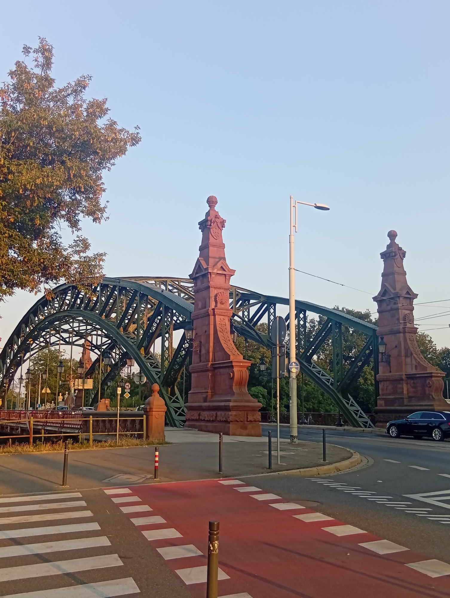 The image shows the Grunwald Bridge in Wrocław, Poland, a historic steel arch bridge spanning the Oder River. In the foreground, a crosswalk with red and white markings is visible, indicating a pedestrian crossing on the road. The bridge itself is a prominent structure, painted green, with intricate metalwork. Two large, ornate reddish-brown stone pillars flank the entrance to the bridge. A dark-colored car is driving on the road to the right of the bridge. Trees with autumn foliage line the background, and the sky is clear and blue, suggesting it is daytime. Streetlights and traffic signs are also visible. The scene depicts a typical urban environment with infrastructure and transportation elements.