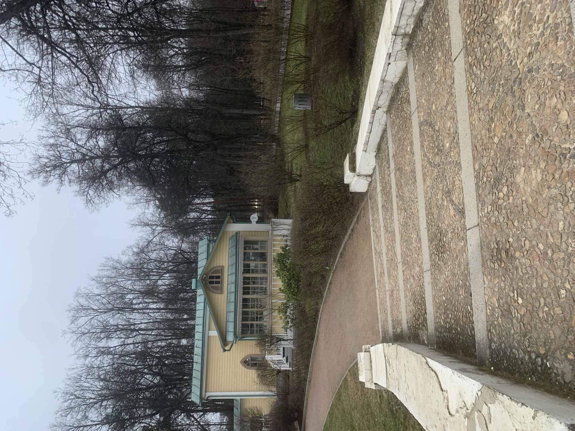 The image depicts a two-story yellow building with green trim, situated in a park-like setting with bare trees and a gray sky. A stone staircase with white railings leads down into the park. A dirt path curves alongside the building. The building's architectural style suggests it might be a historical structure. The location is Moscow, Russia.